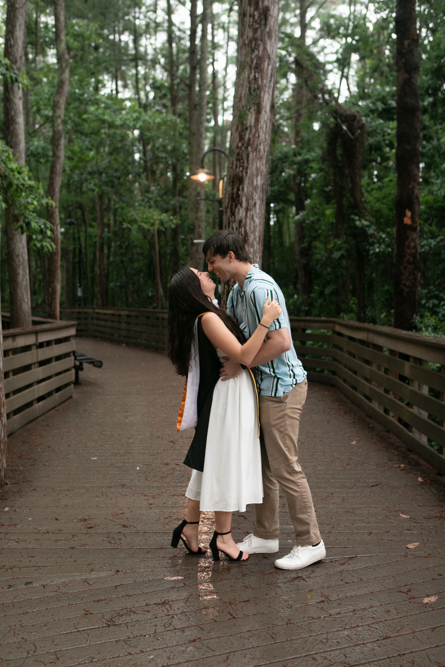 Young couple shares a romantic kiss on a rain-wet wooden walkway surrounded by tall pine trees.