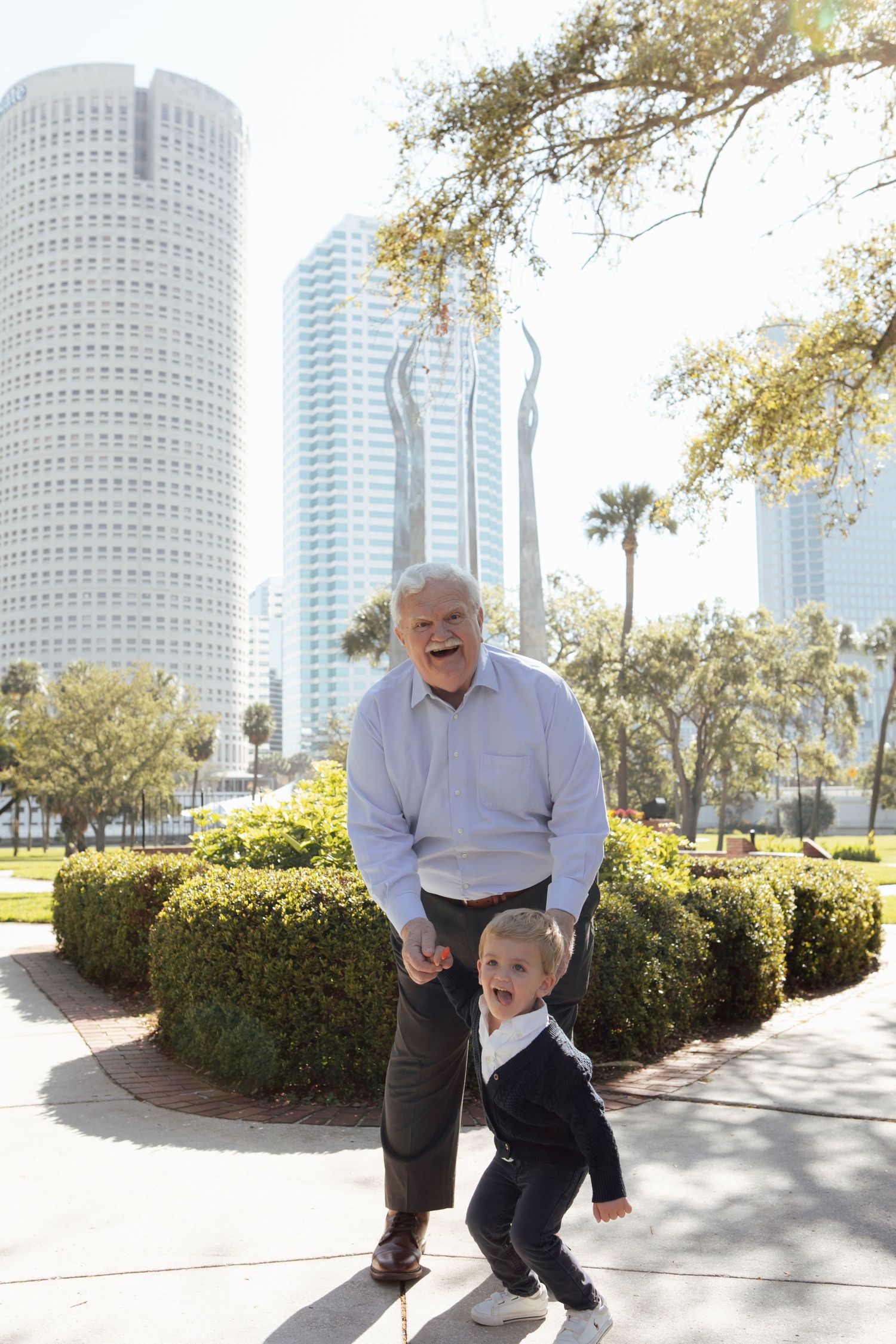 Laughing grandfather and excited toddler boy pose together in a sunny city park with tall skyscrapers behind them.