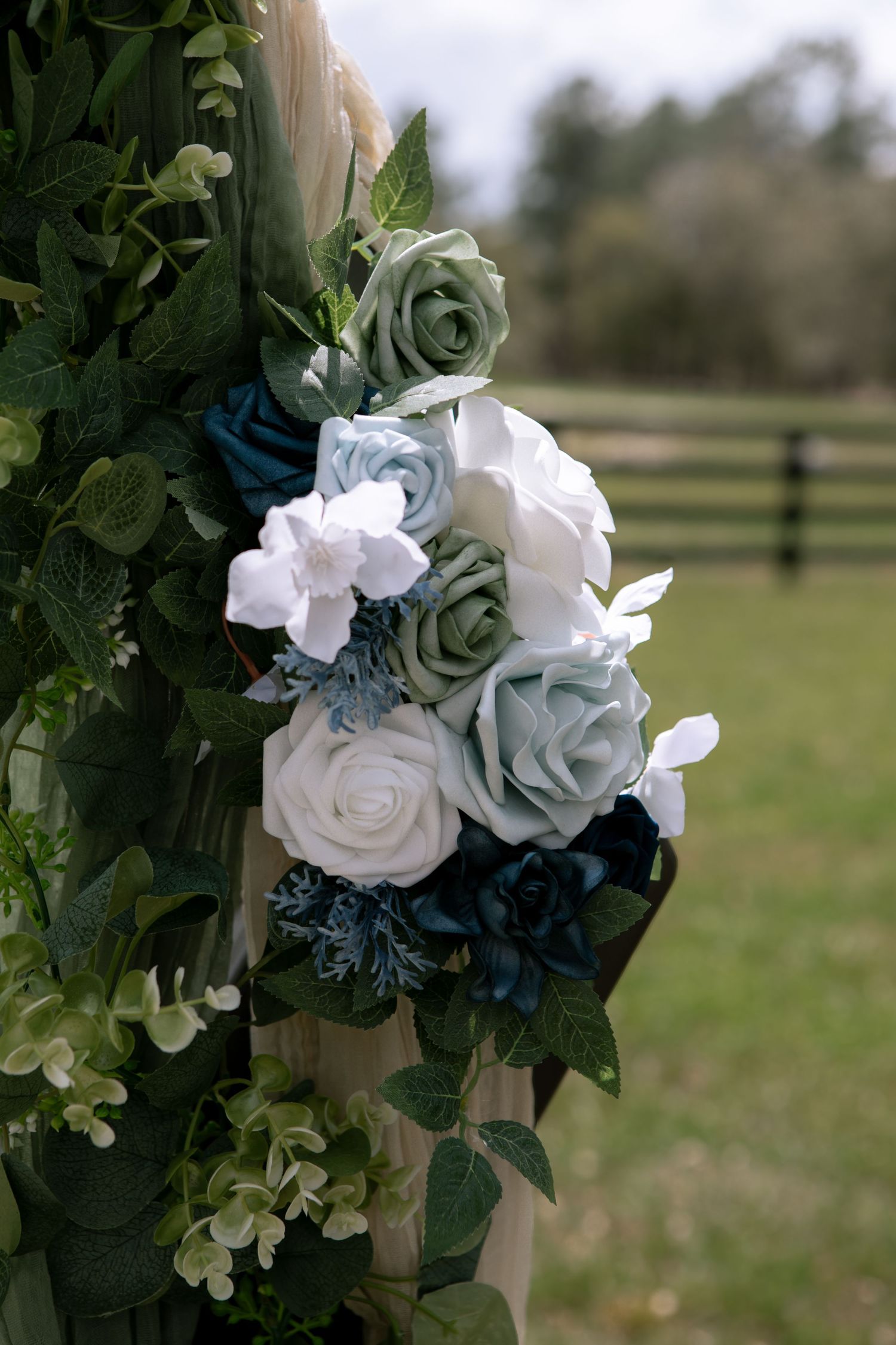 Elegant wedding arch floral arrangement featuring white, sage green, and navy blue roses with lush greenery outdoors.