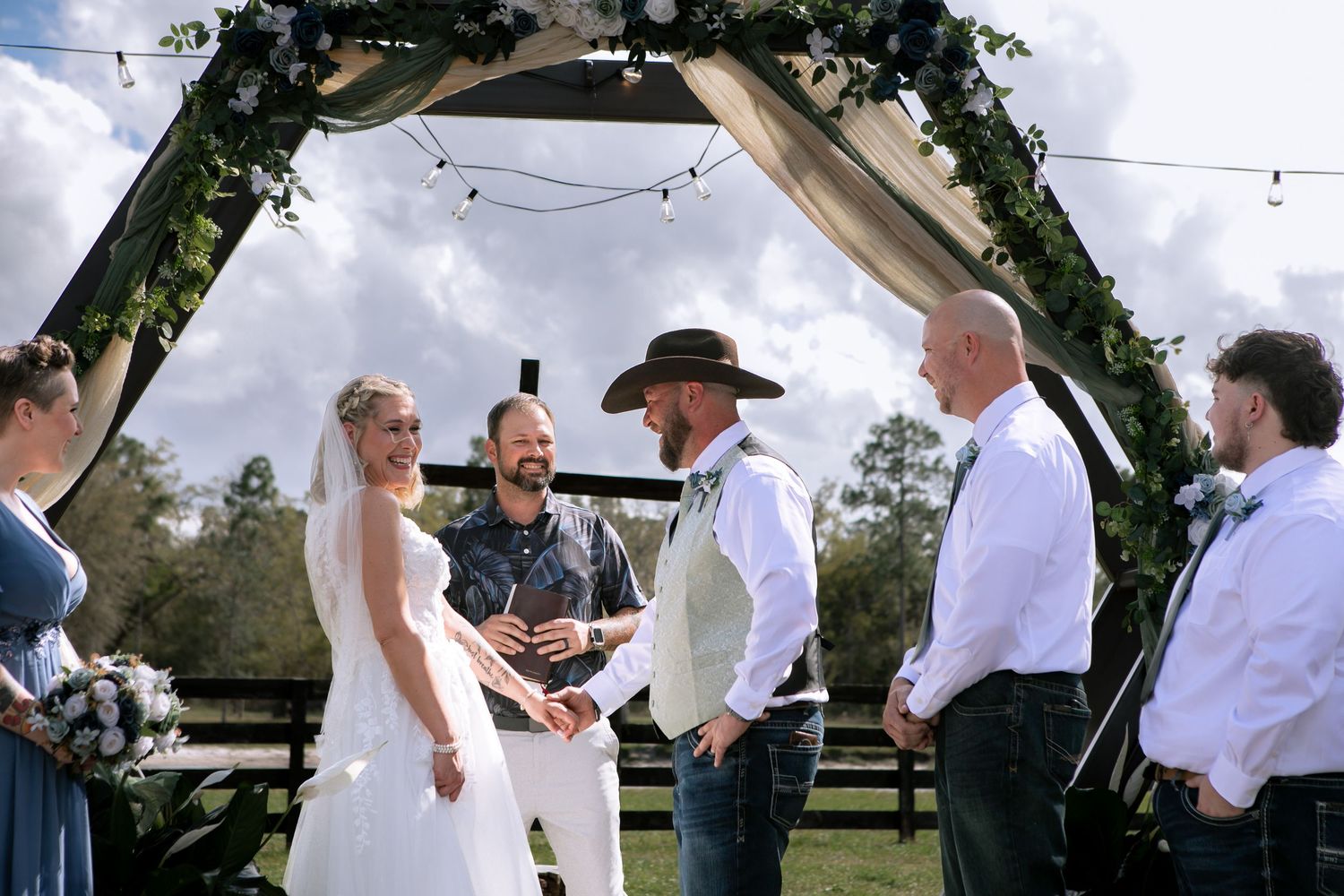 Outdoor wedding ceremony under floral arch with bride in white gown, groom in cowboy hat, and officiant on green lawn.