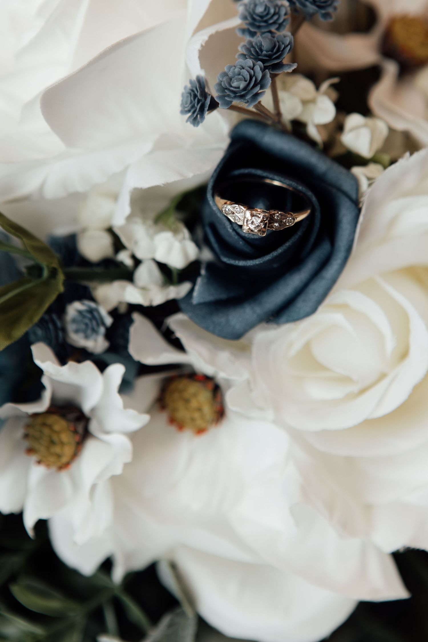Gold diamond engagement ring nestled in a bridal bouquet of white and navy blue roses.