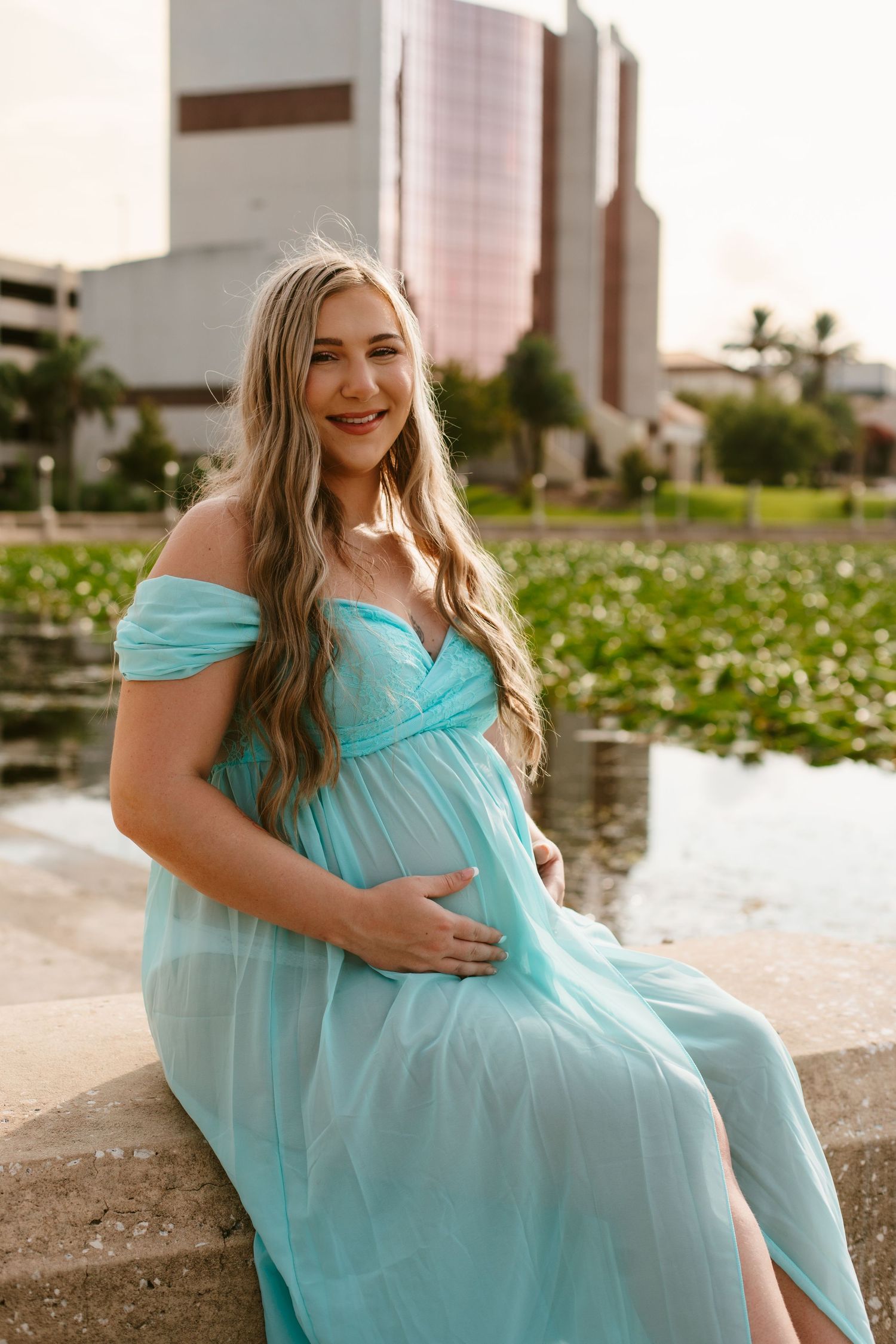 Pregnant woman in teal off-shoulder gown sitting by a pond, smiling warmly during a maternity photo session.