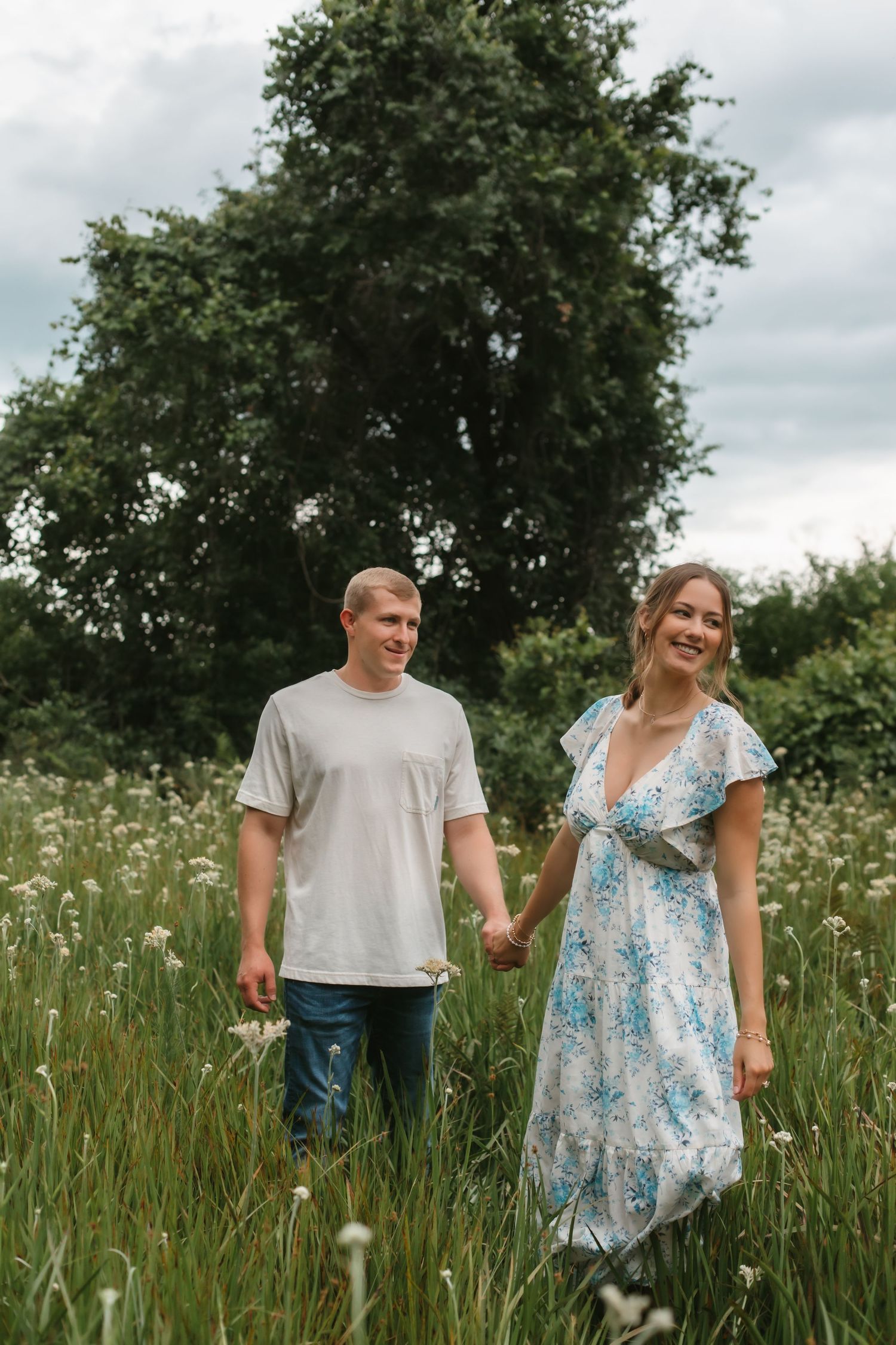 A smiling couple holds hands while walking through a lush green meadow with wildflowers on a cloudy day.