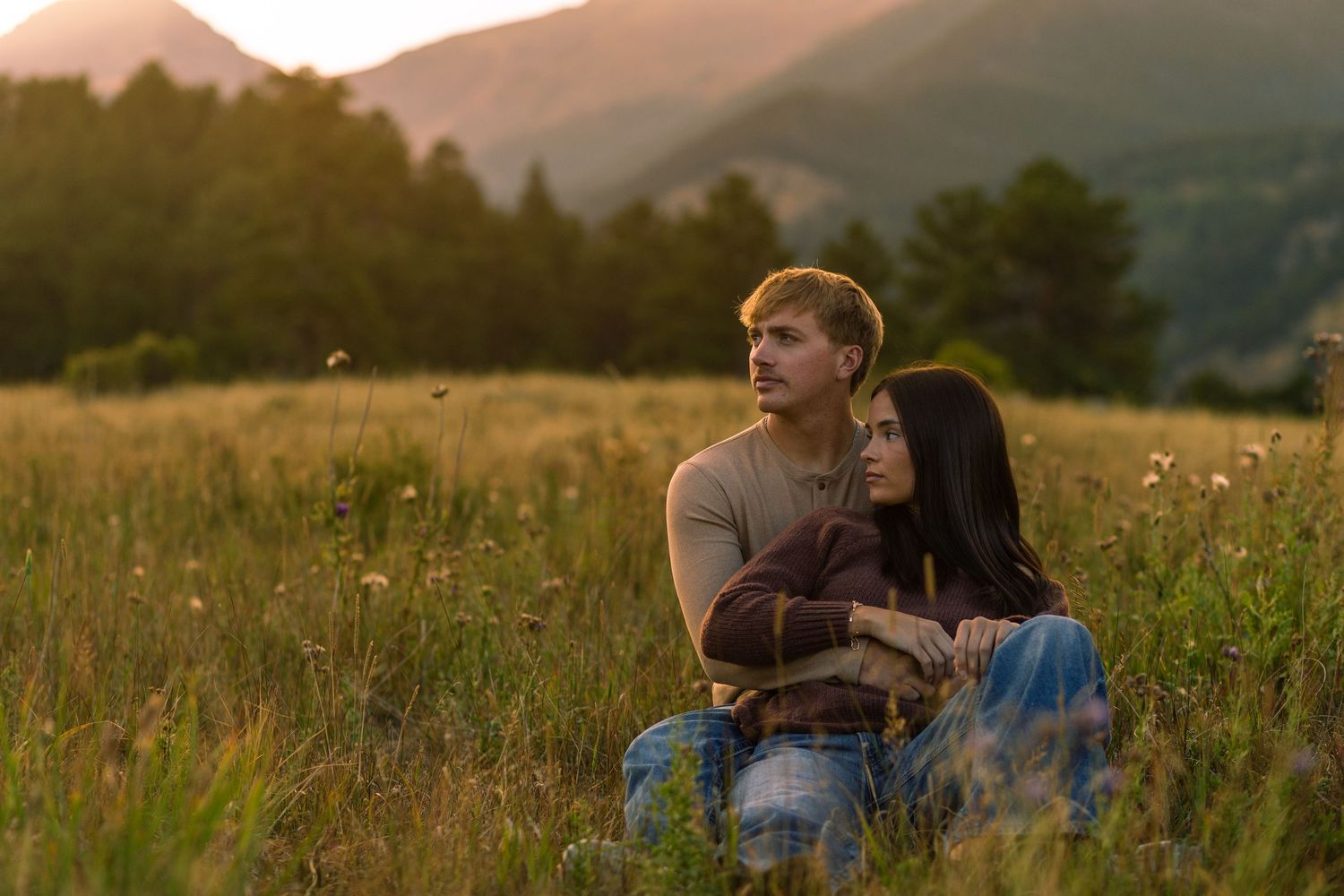 A couple sits together in a golden meadow at sunset with mountains and forest in the background.