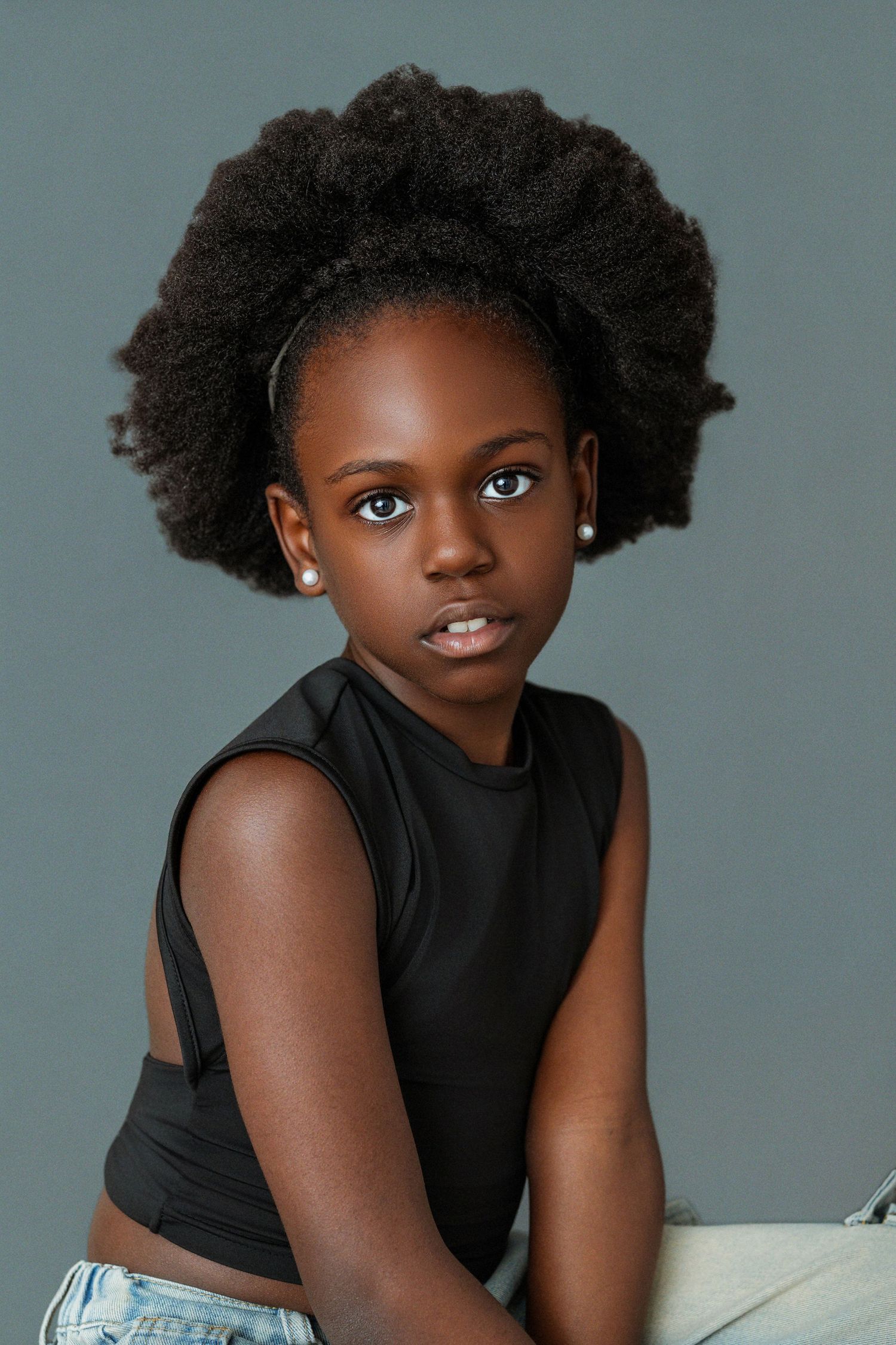 Young Black girl with natural afro hair wearing a black sleeveless top and jeans, posing confidently against a gray background.