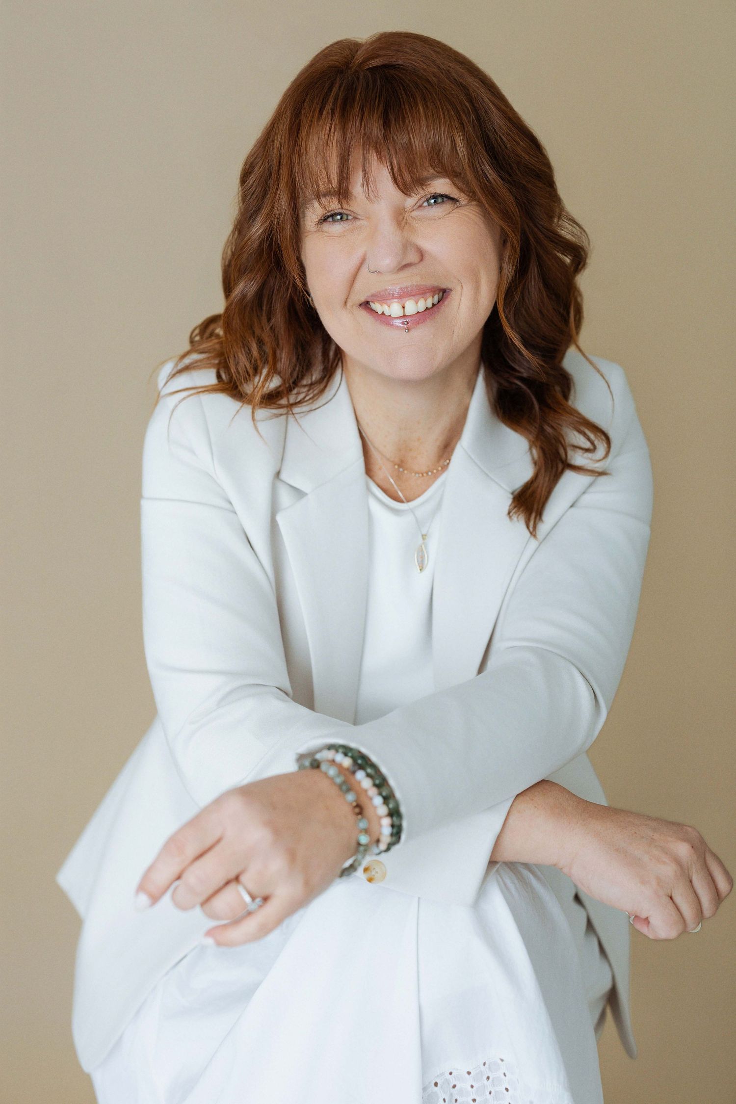 Smiling woman with auburn hair wearing a white blazer and beaded bracelets, posing confidently against a beige background.