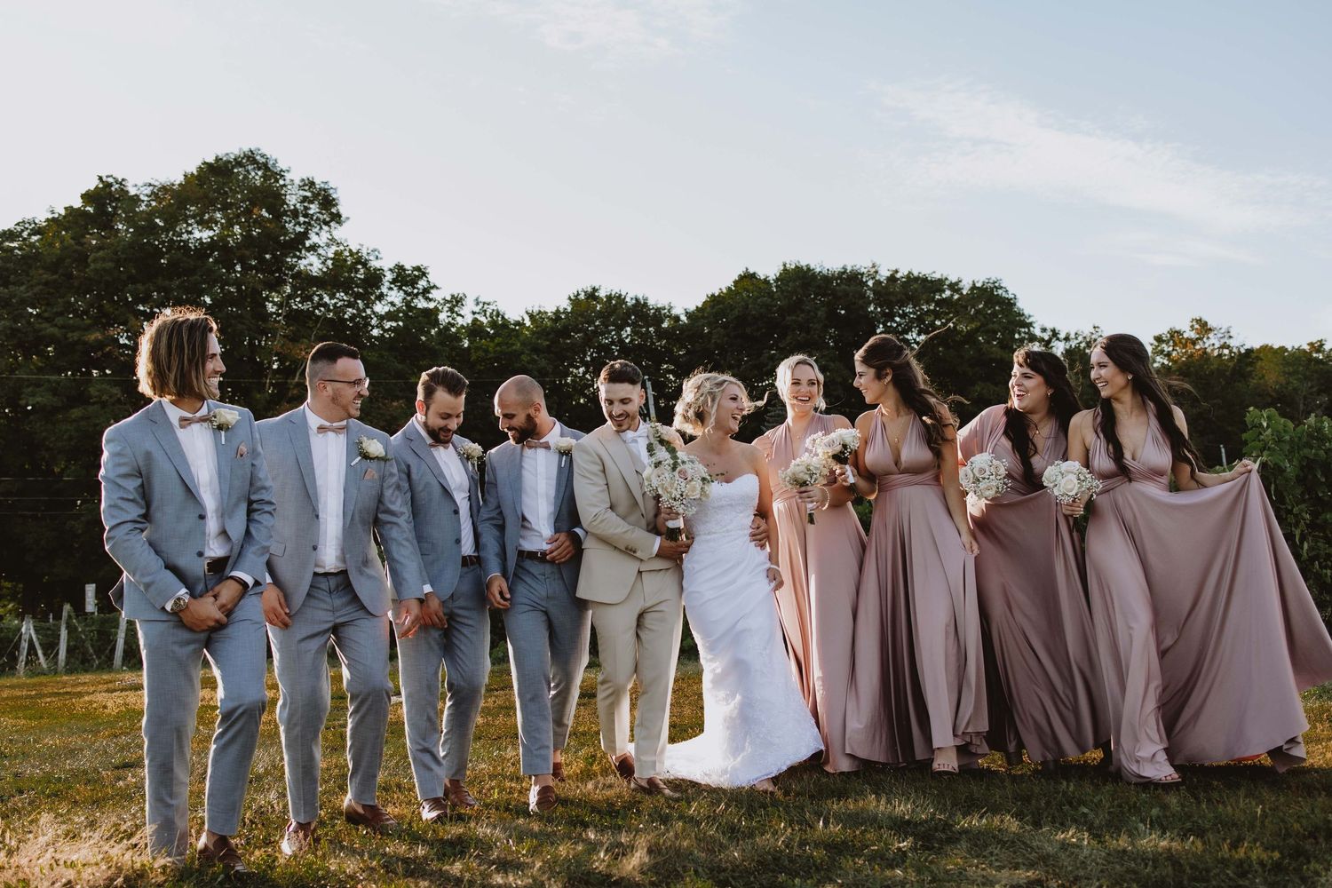 Wedding party in light blue suits and mauve dresses posing outdoors on grassy field.