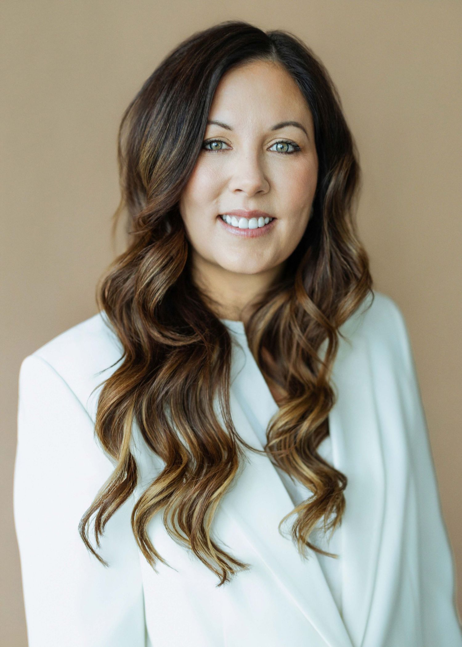 Professional headshot of a smiling woman with long wavy brunette hair wearing a white blazer against a beige background.