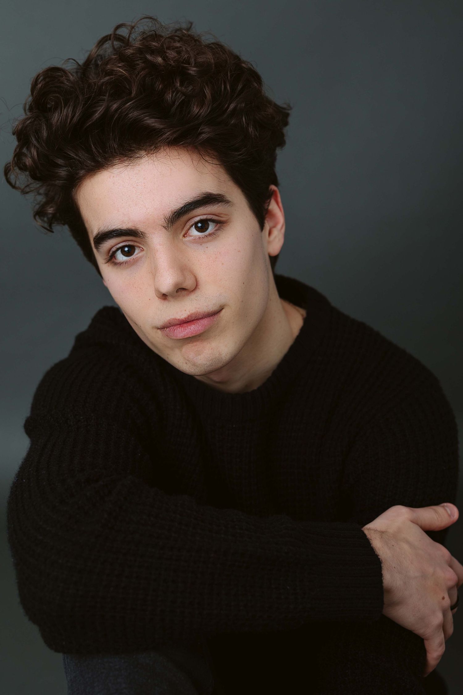 Portrait of a young person with dark curly hair wearing a black sweater against a dark background.