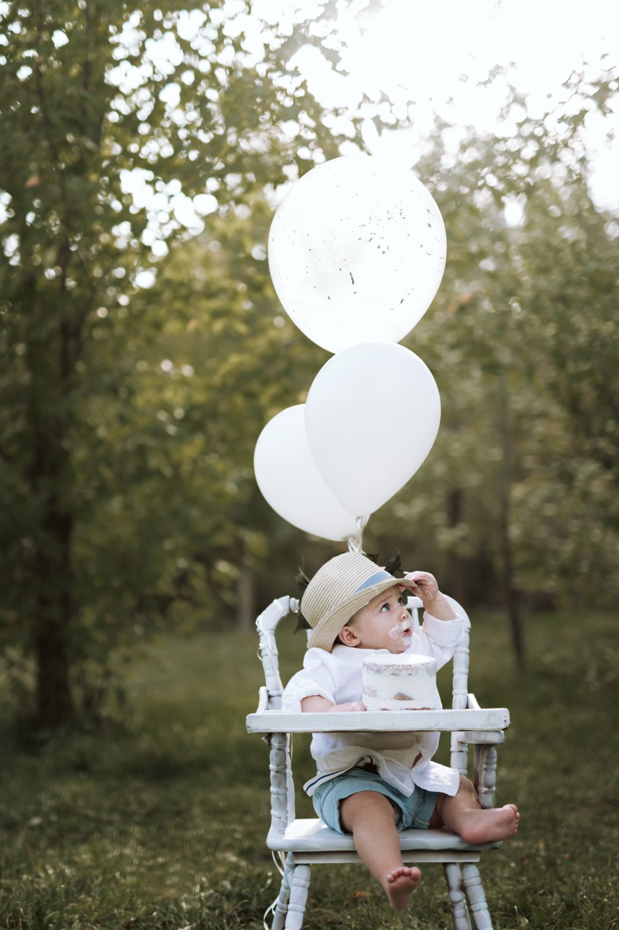 Baby sitting in white high chair with heart shaped balloons in outdoor garden setting.