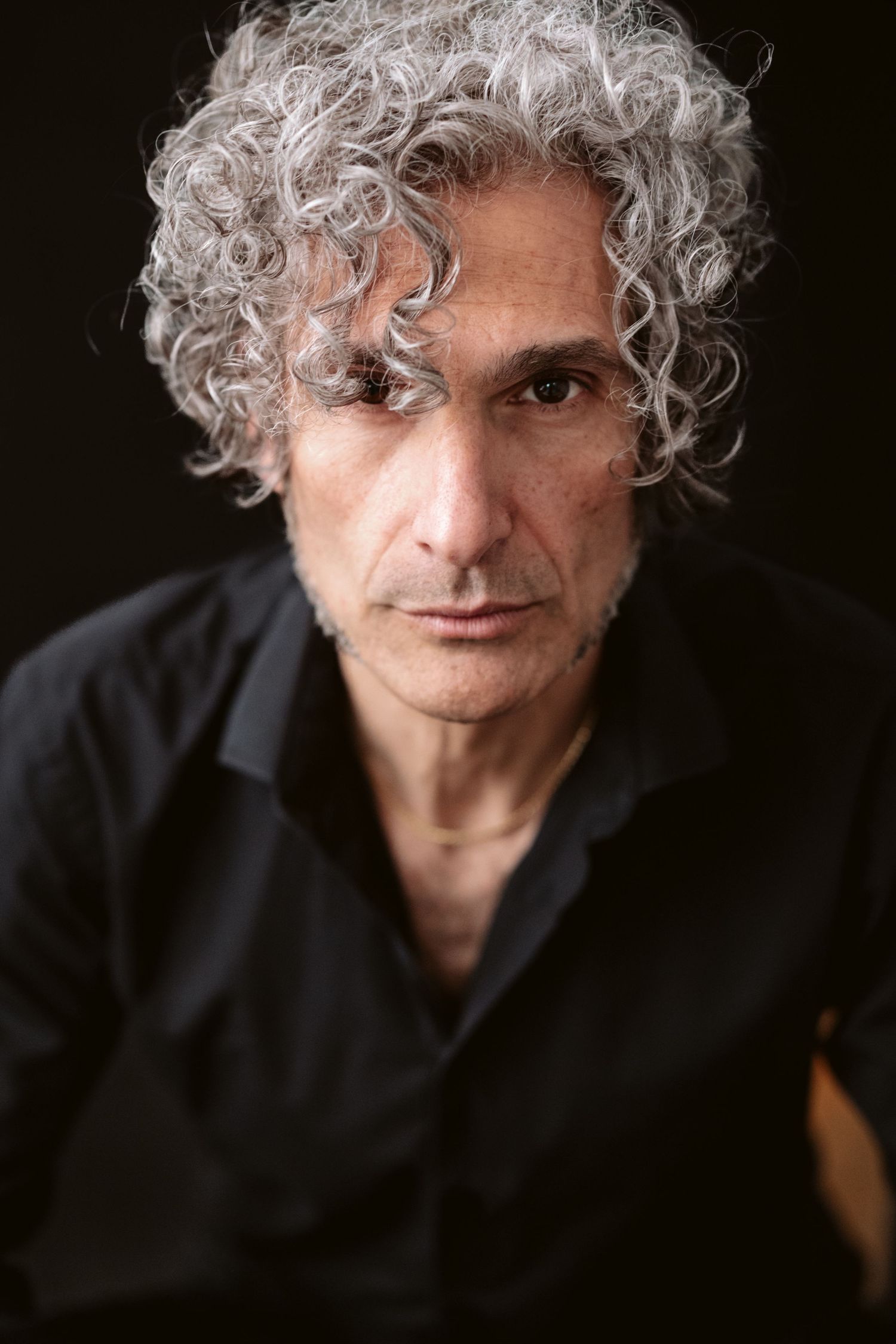 Dramatic portrait series of person with curly gray hair wearing black shirt against dark background.
