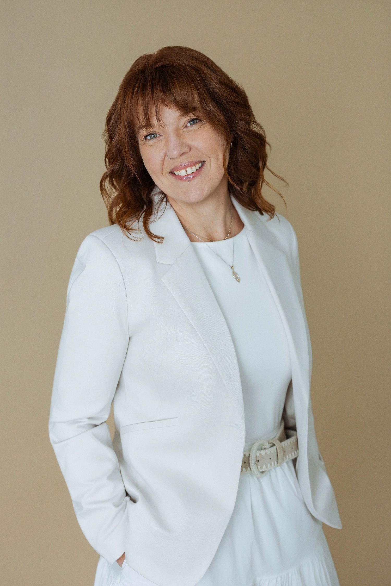 Professional woman in white blazer and dress smiling confidently against a neutral beige background.