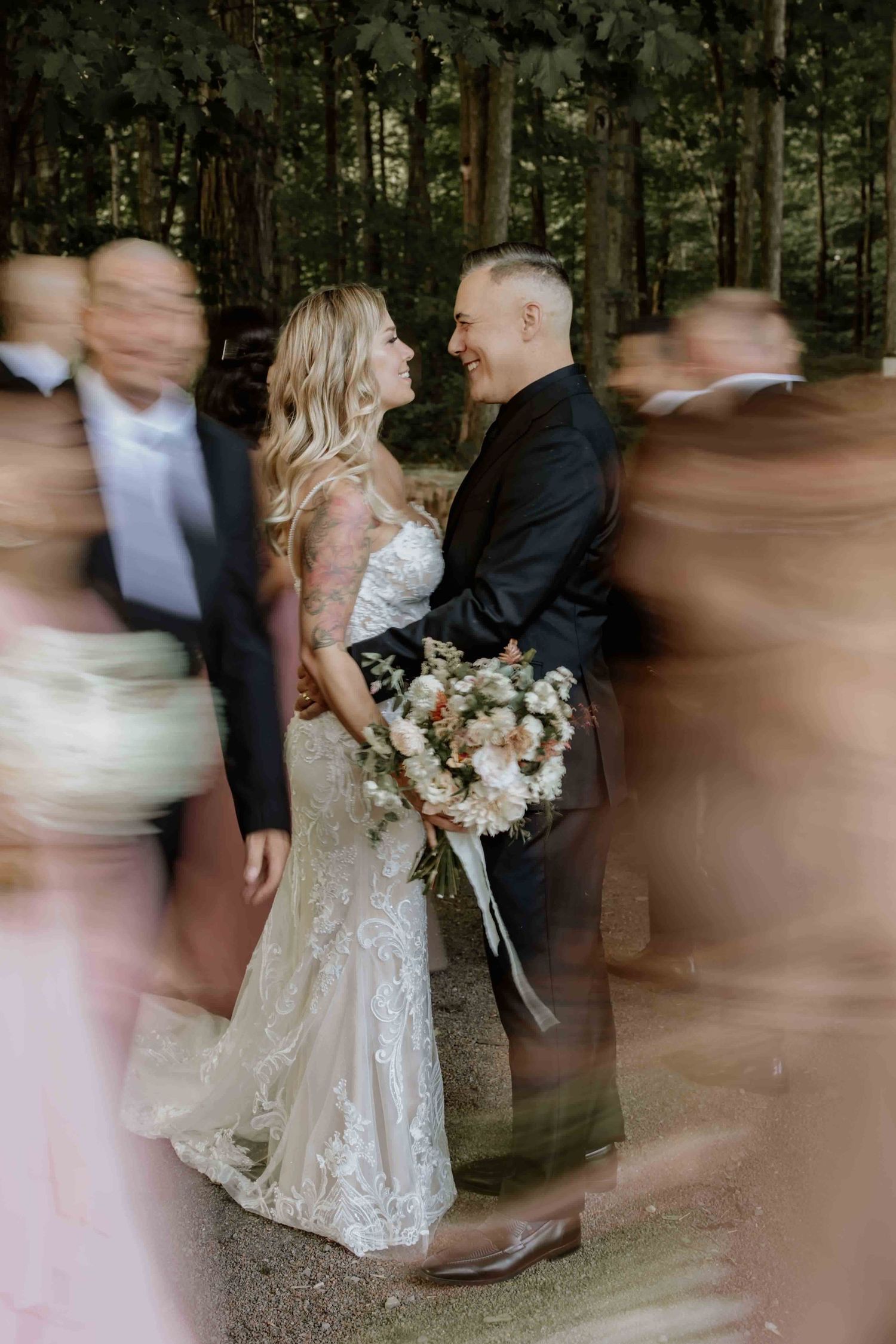 Artistic wedding photo with motion blur of guests walking past while couple stands still in forest setting with bouquet.