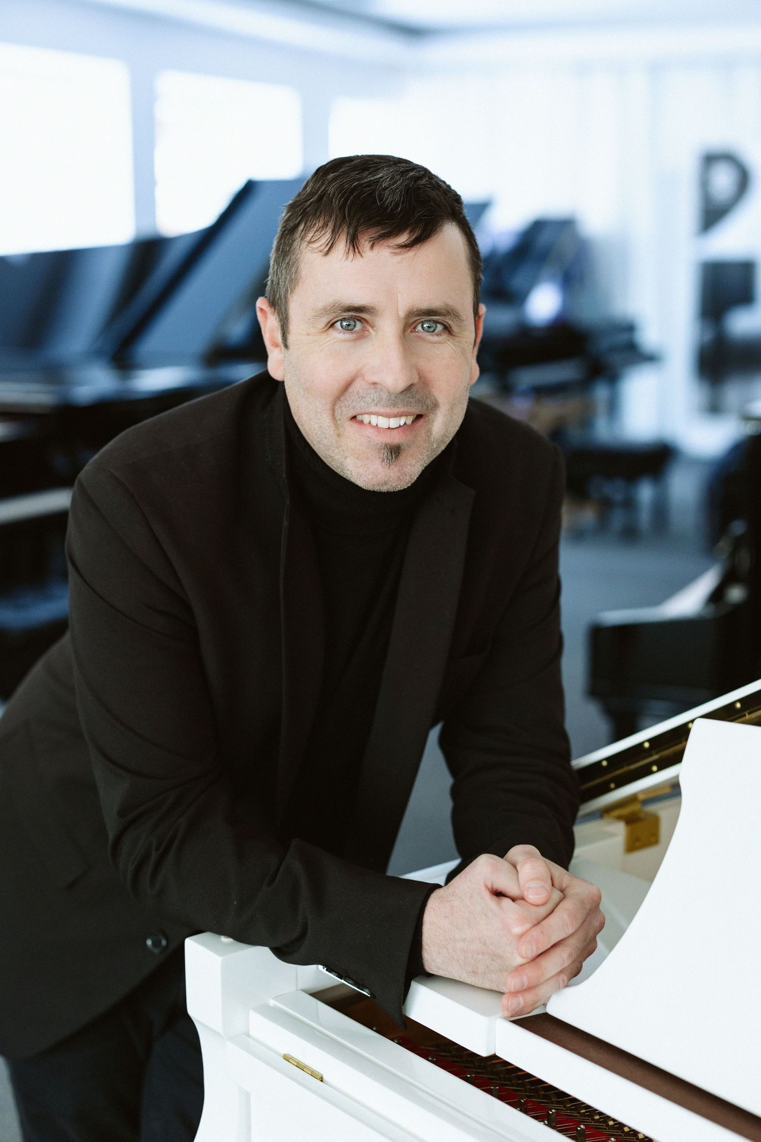 Professional headshot of someone in a black sweater leaning on a white piano in a modern setting.