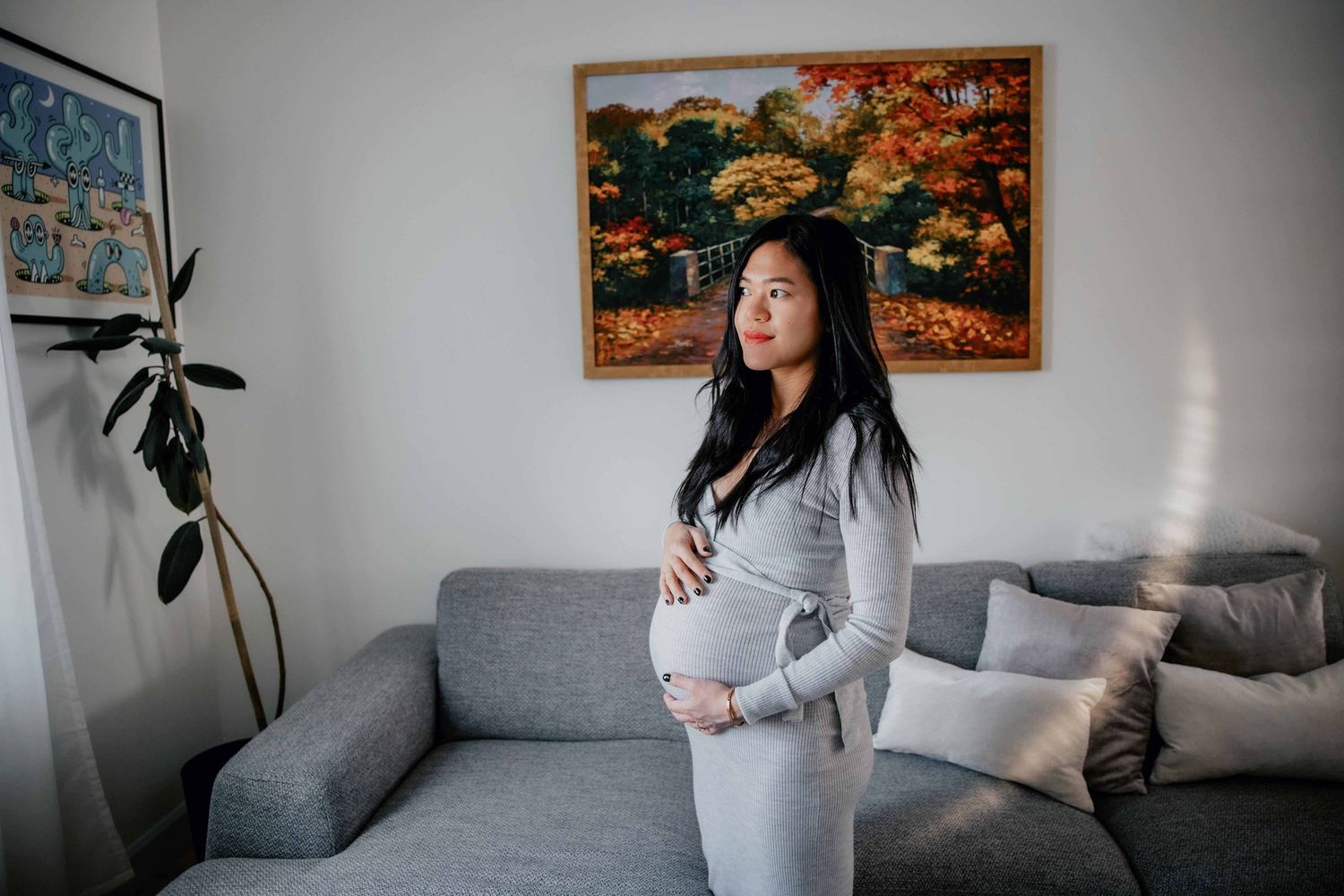Pregnant woman in grey dress standing near artwork in modern living room.
