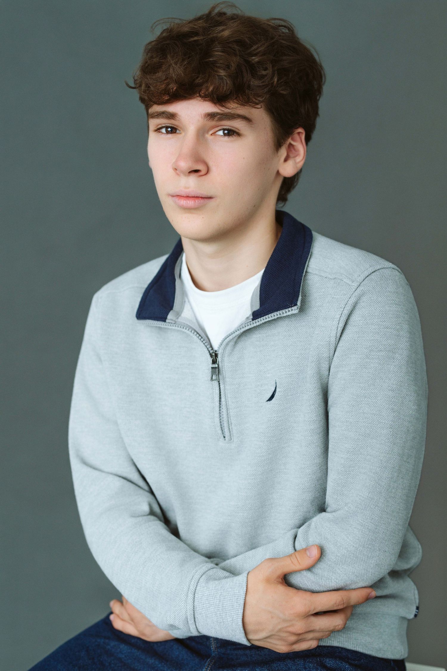 Young teen boy with curly brown hair wearing a gray Nautica quarter-zip sweatshirt, sitting with arms crossed.