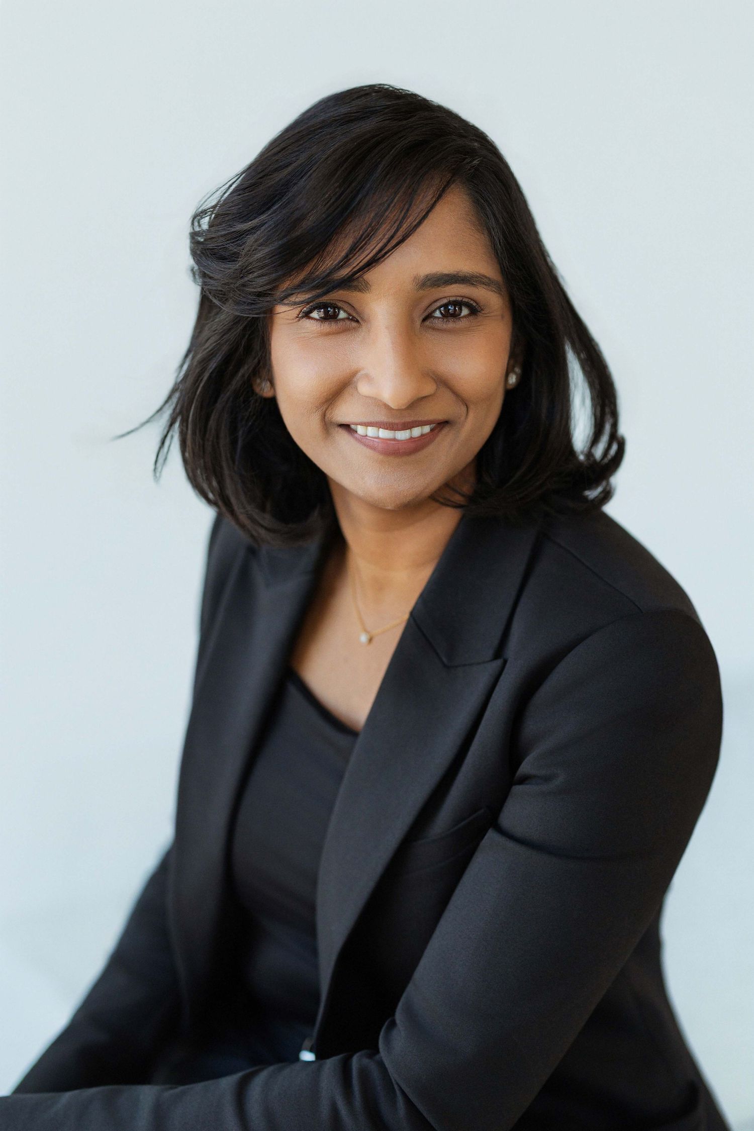 Professional headshot of a smiling South Asian woman in a black blazer against a light background.
