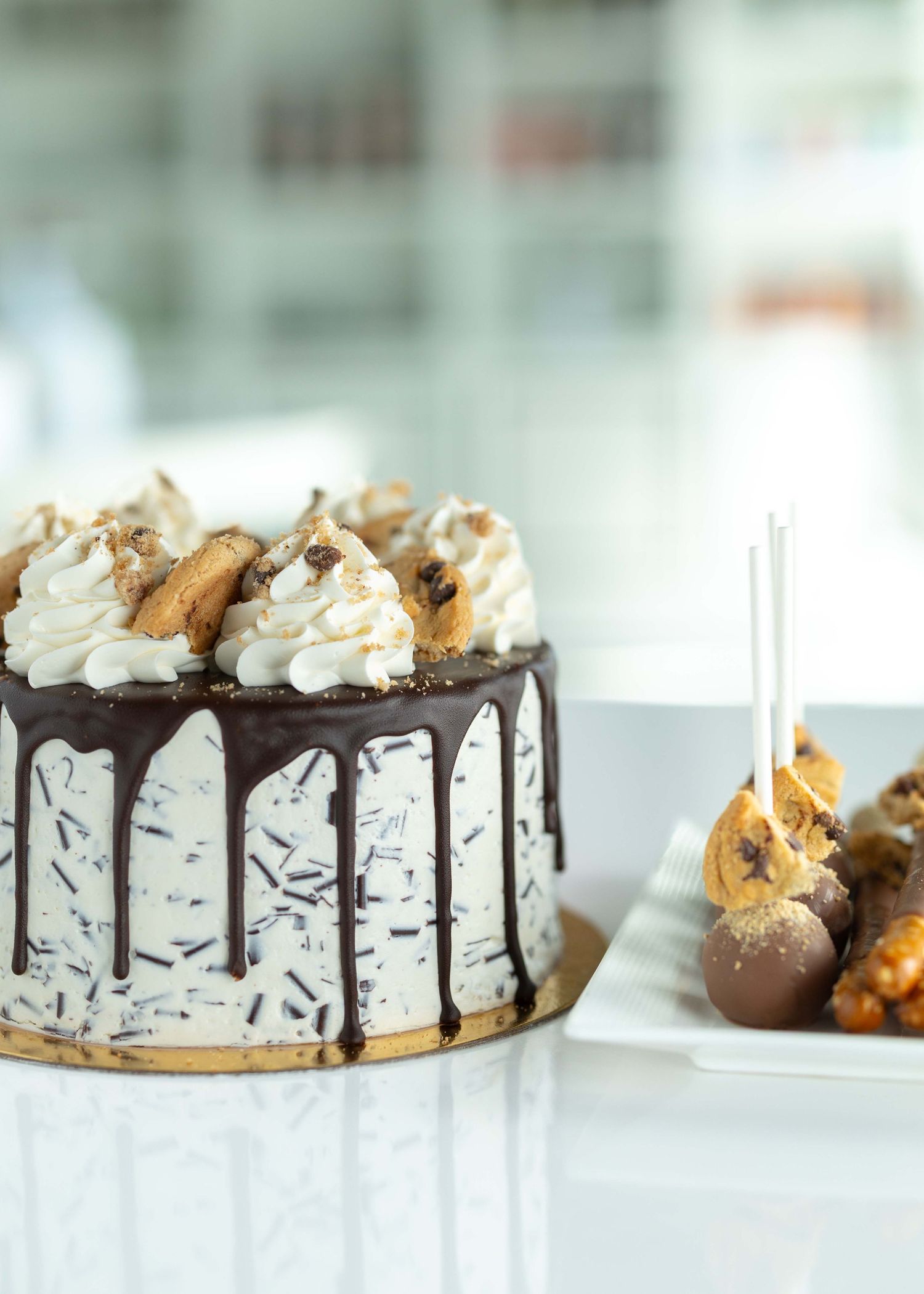 A decadent chocolate drip cake topped with whipped cream and cookie pieces on a white cake stand.