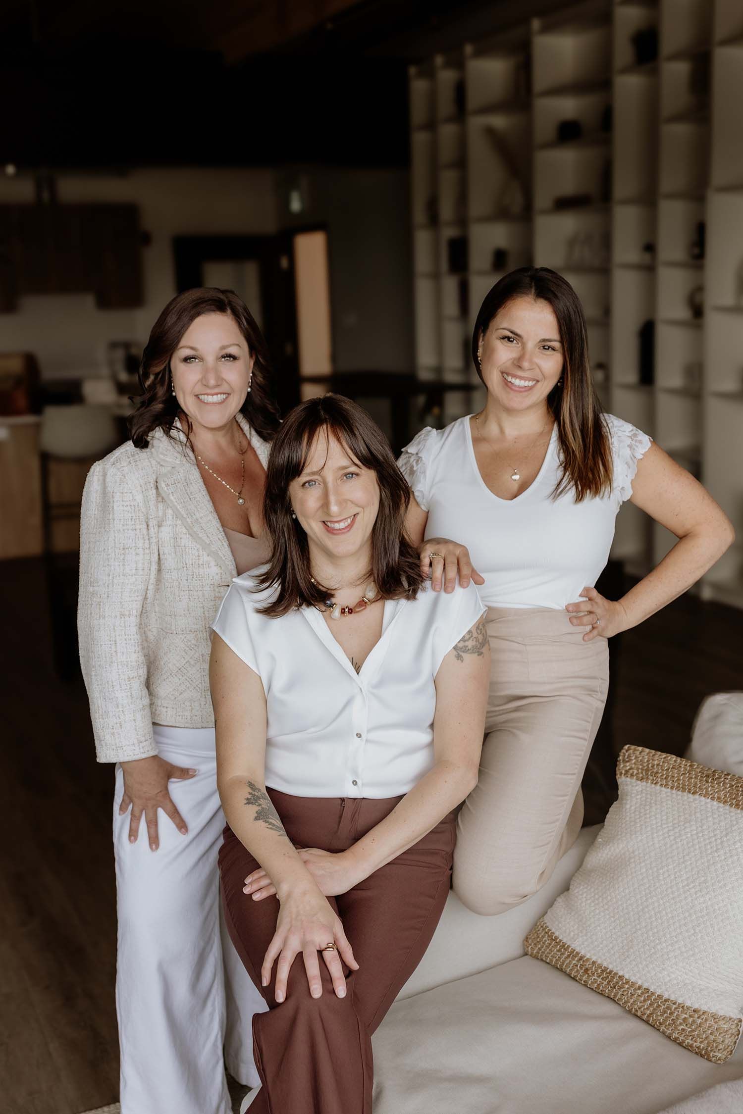 Three smiling women pose together in a modern interior, wearing neutral tones of white and brown.