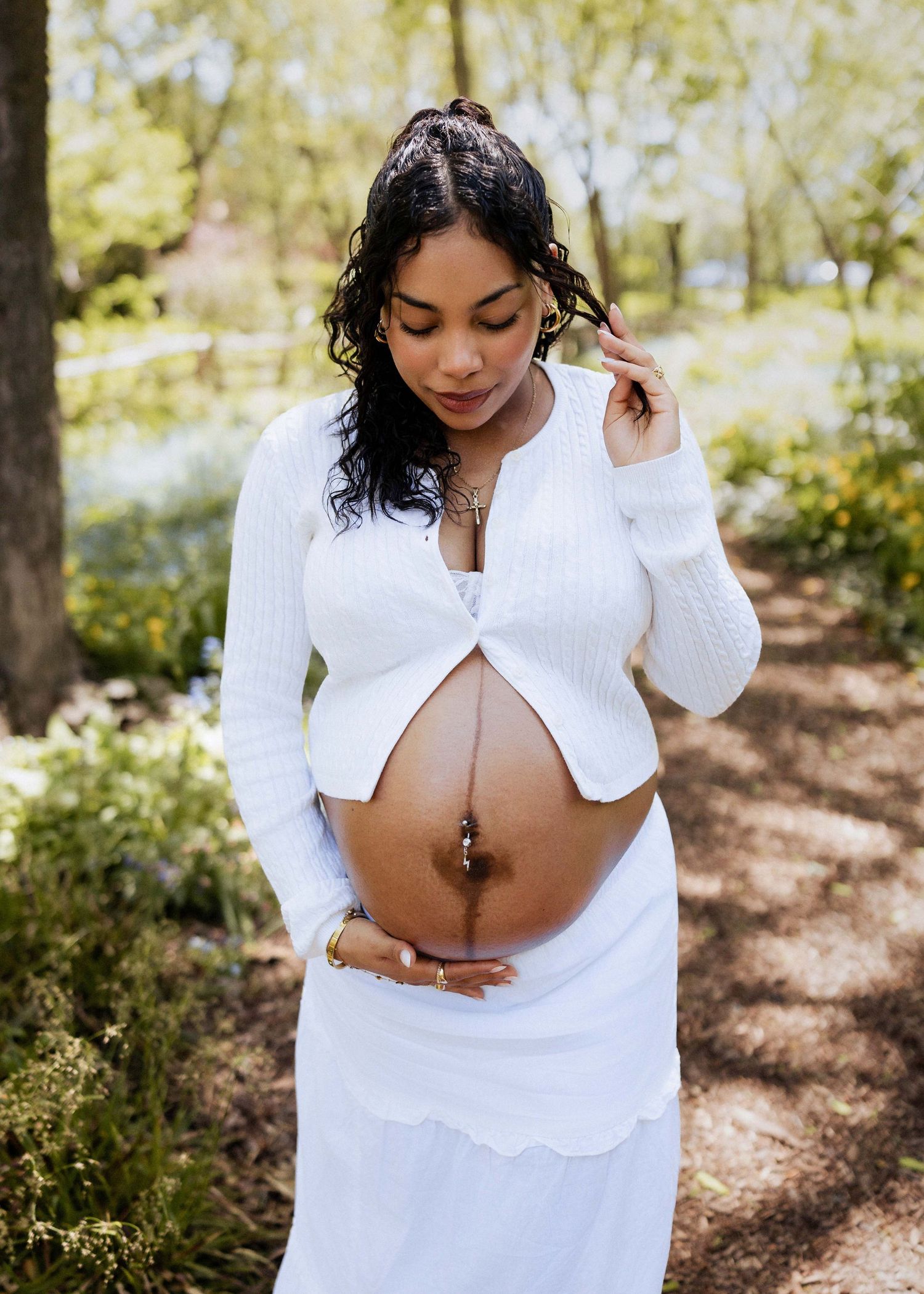 Pregnant woman in white outfit outdoors, cradling bare baby bump, surrounded by lush green trees.