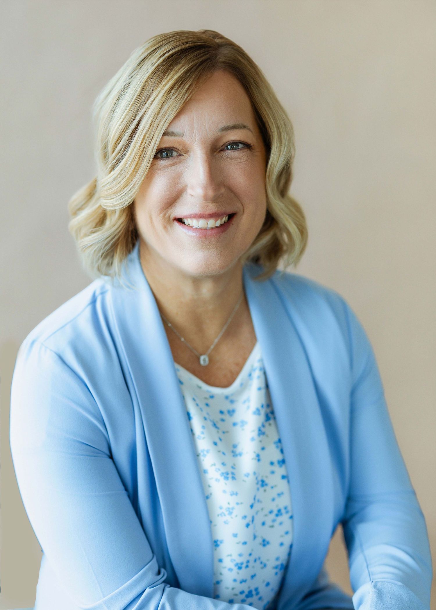 Smiling blonde woman in light blue cardigan and floral top poses confidently against a neutral background.