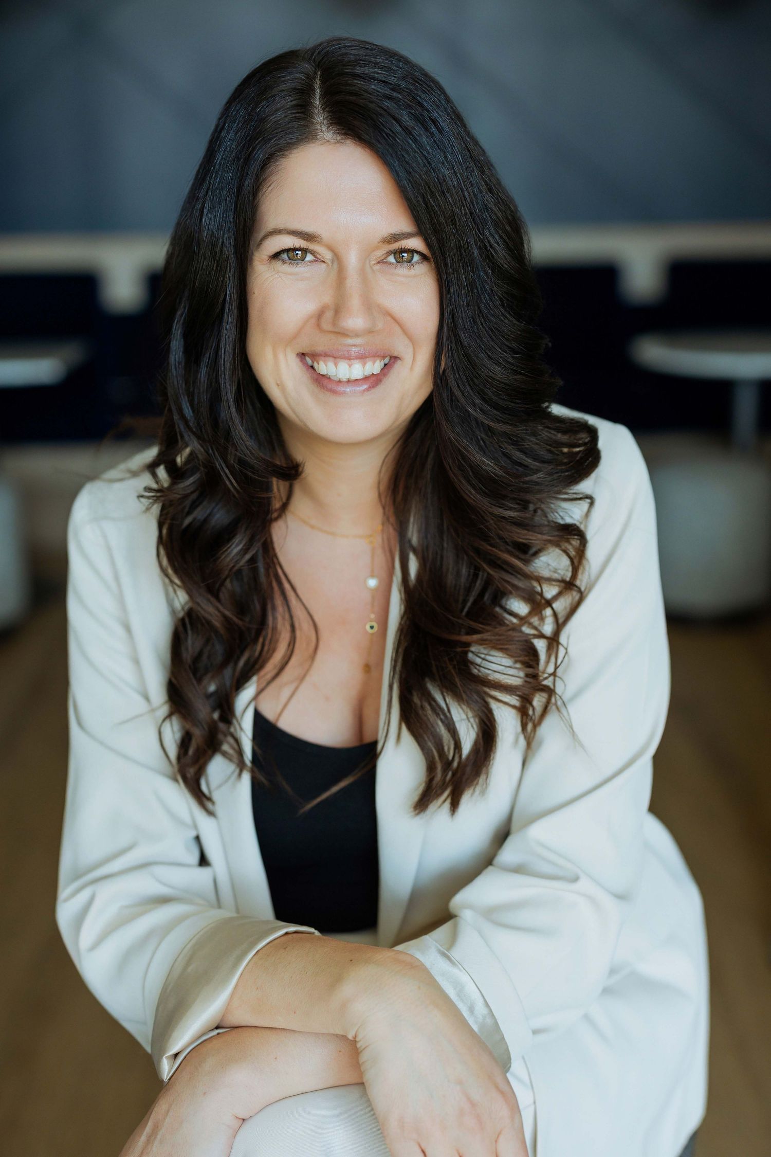 Smiling woman with dark wavy hair wearing a white blazer and black top, seated in a modern office setting.
