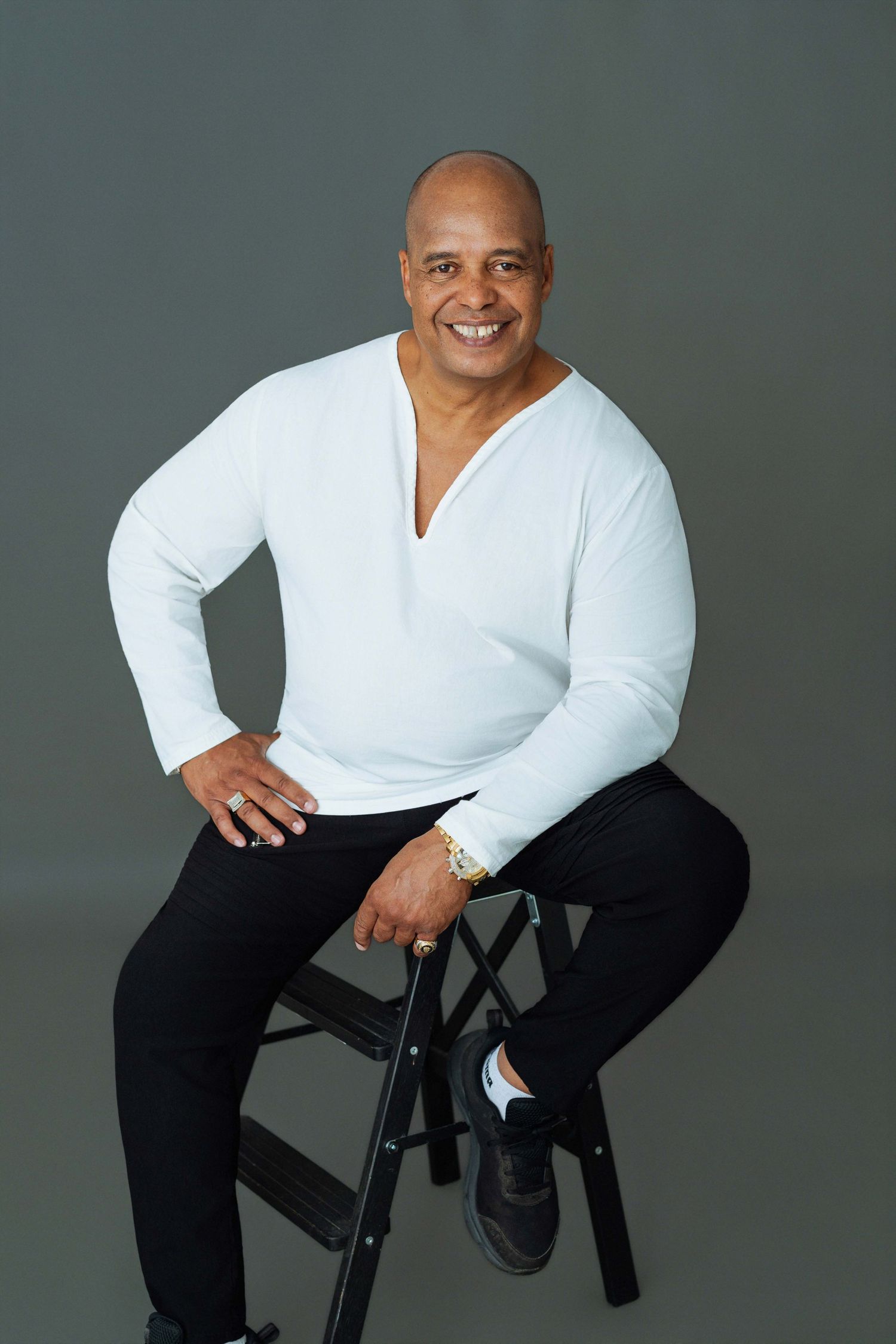 Smiling bald man sitting on black stool wearing white long-sleeve shirt and black pants in studio photo.