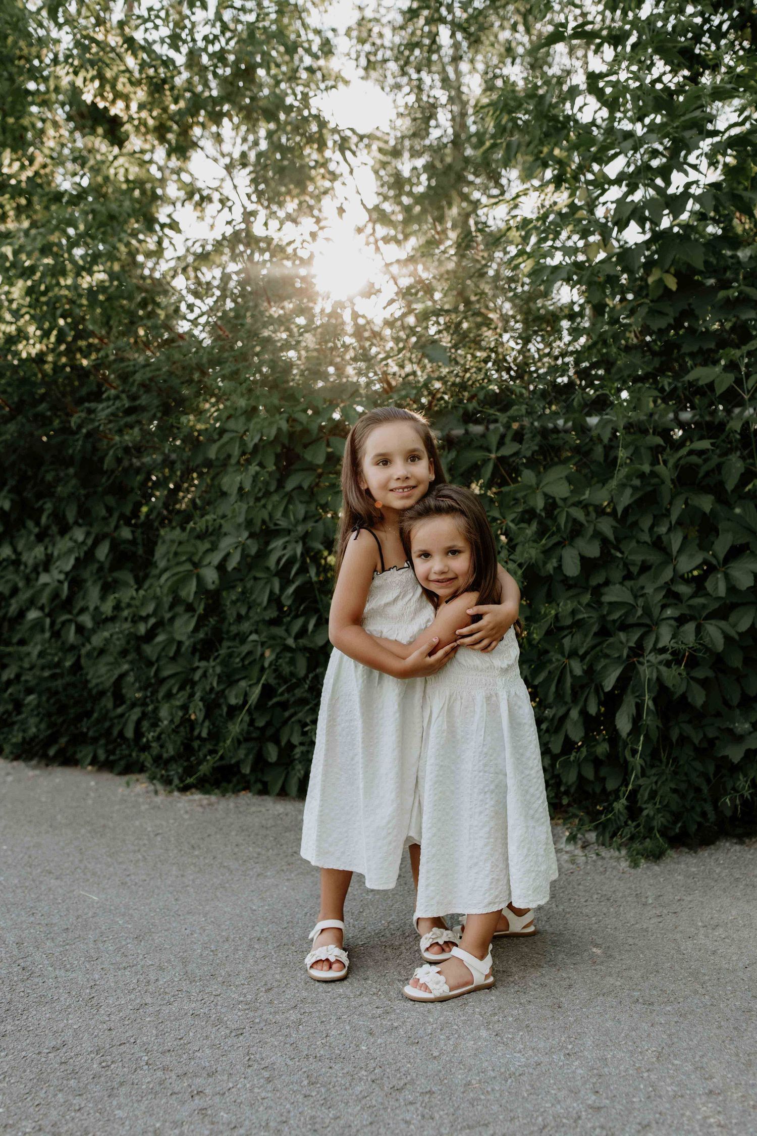 Two young children in white dresses hugging outdoors during sunset.