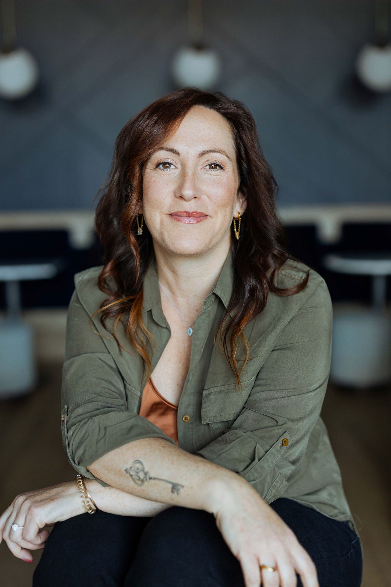 Woman with auburn hair in olive shirt, arms crossed, smiling, with a key tattoo on forearm, seated indoors.