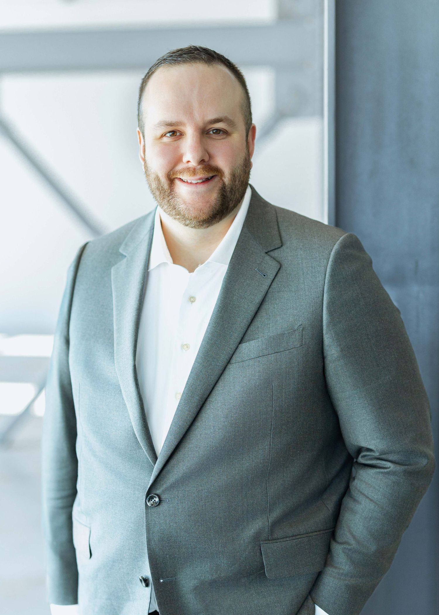 Smiling bearded man in gray suit poses confidently indoors with modern blue-toned background.