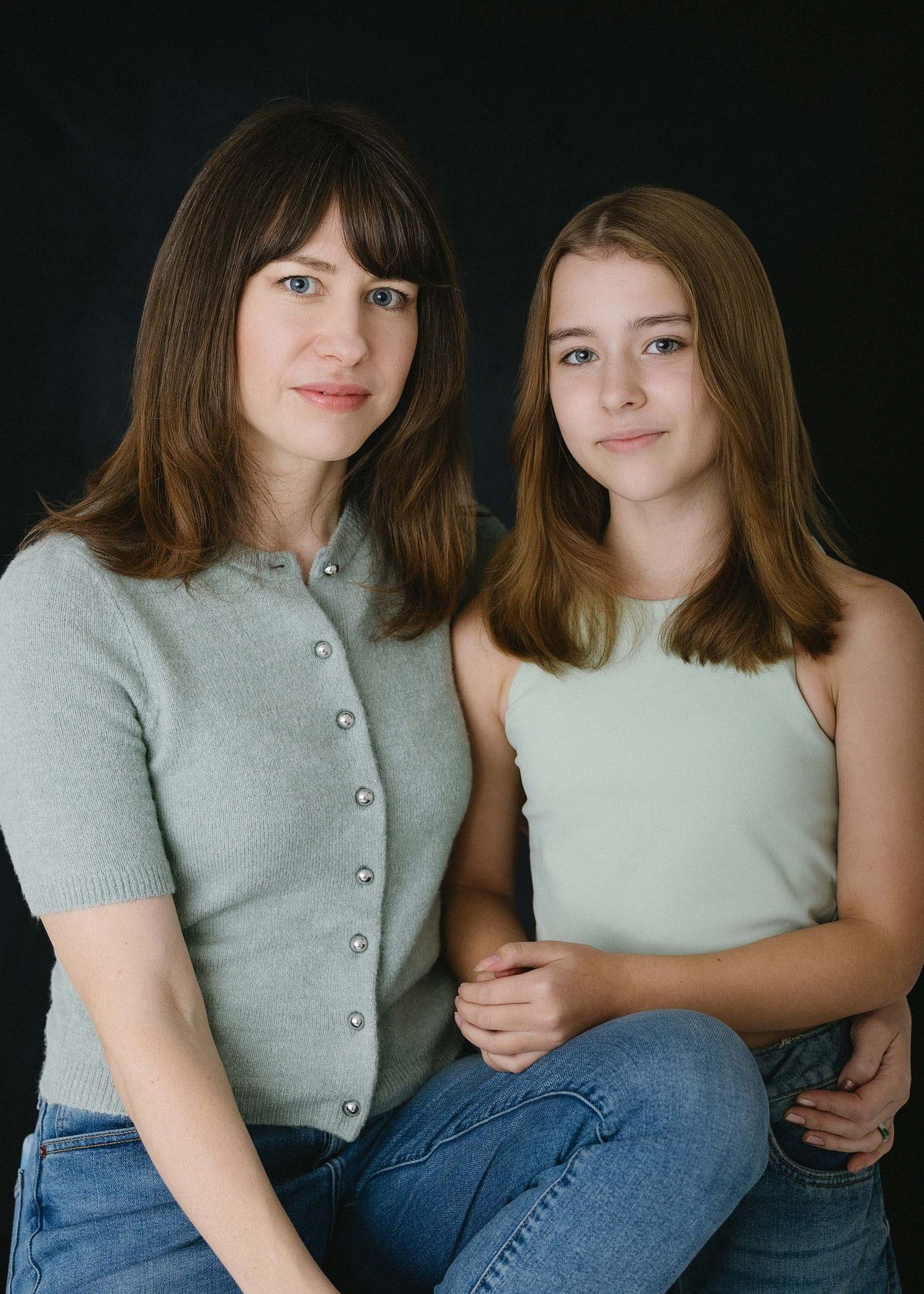 Two people in casual sweaters pose together against dark background in series of intimate portraits.