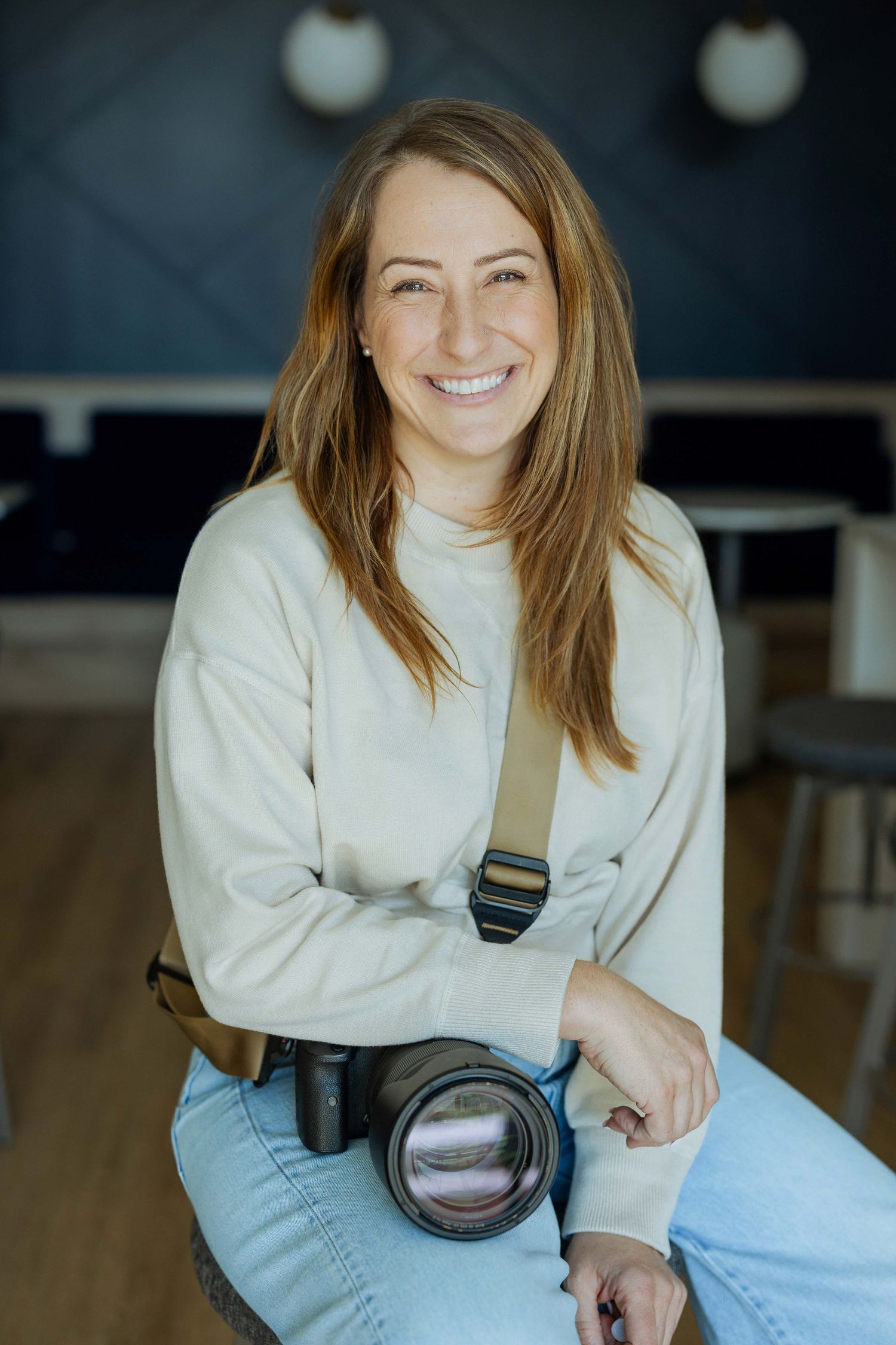 Smiling woman photographer sitting with DSLR camera on strap wearing cream sweater and jeans in modern studio space.