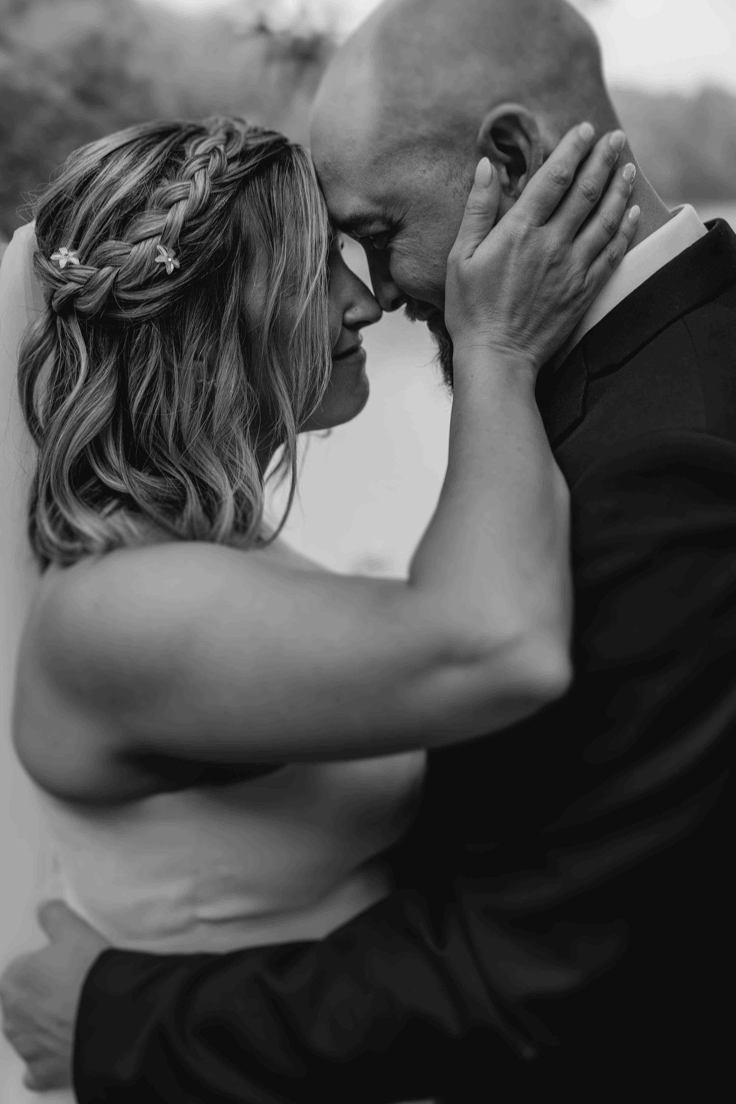 Black and white intimate wedding photo showing tender moment between couple with bride's braided hair and groom in suit.