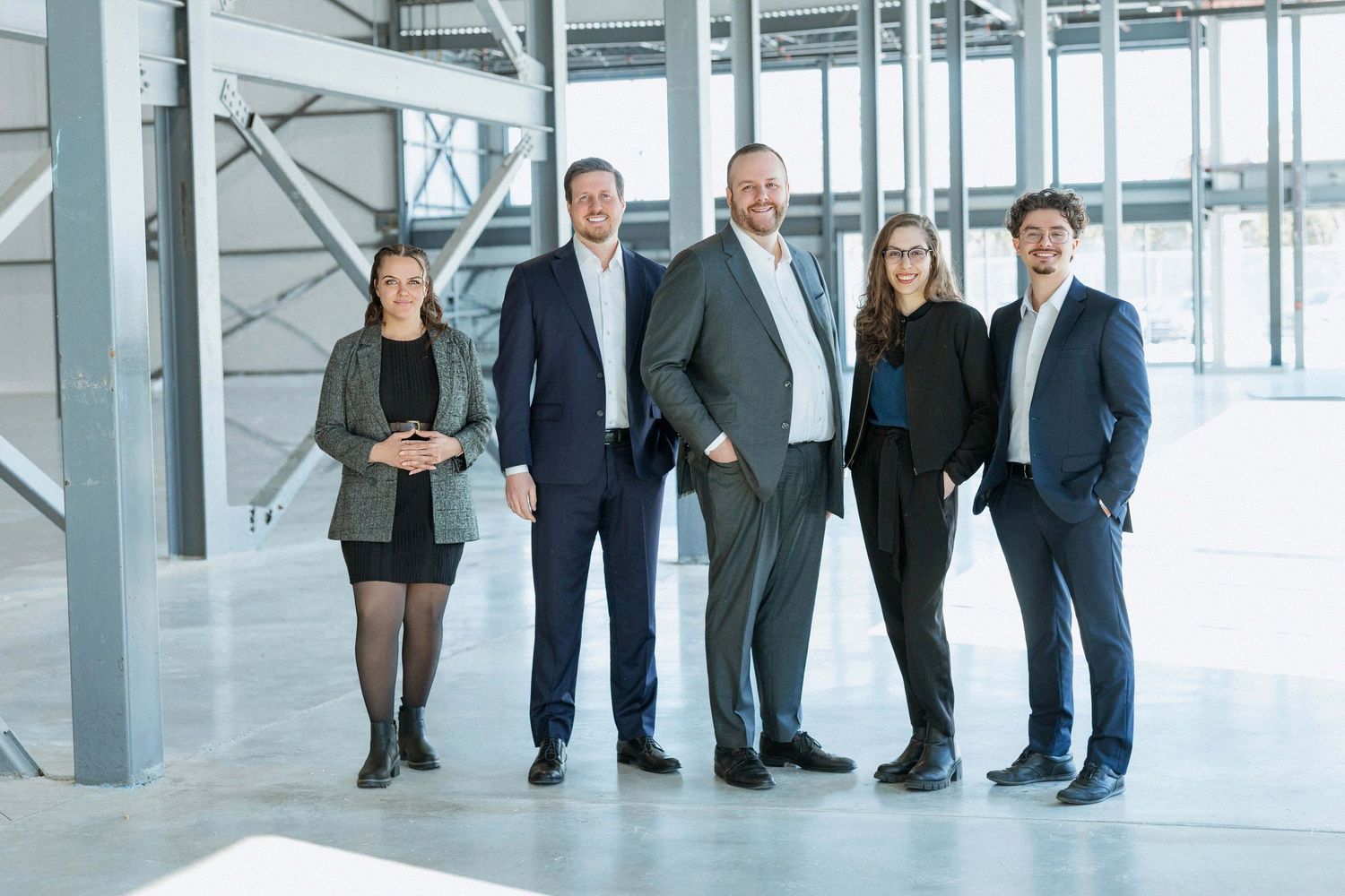 Five business professionals smiling together in a modern industrial office space with large windows.