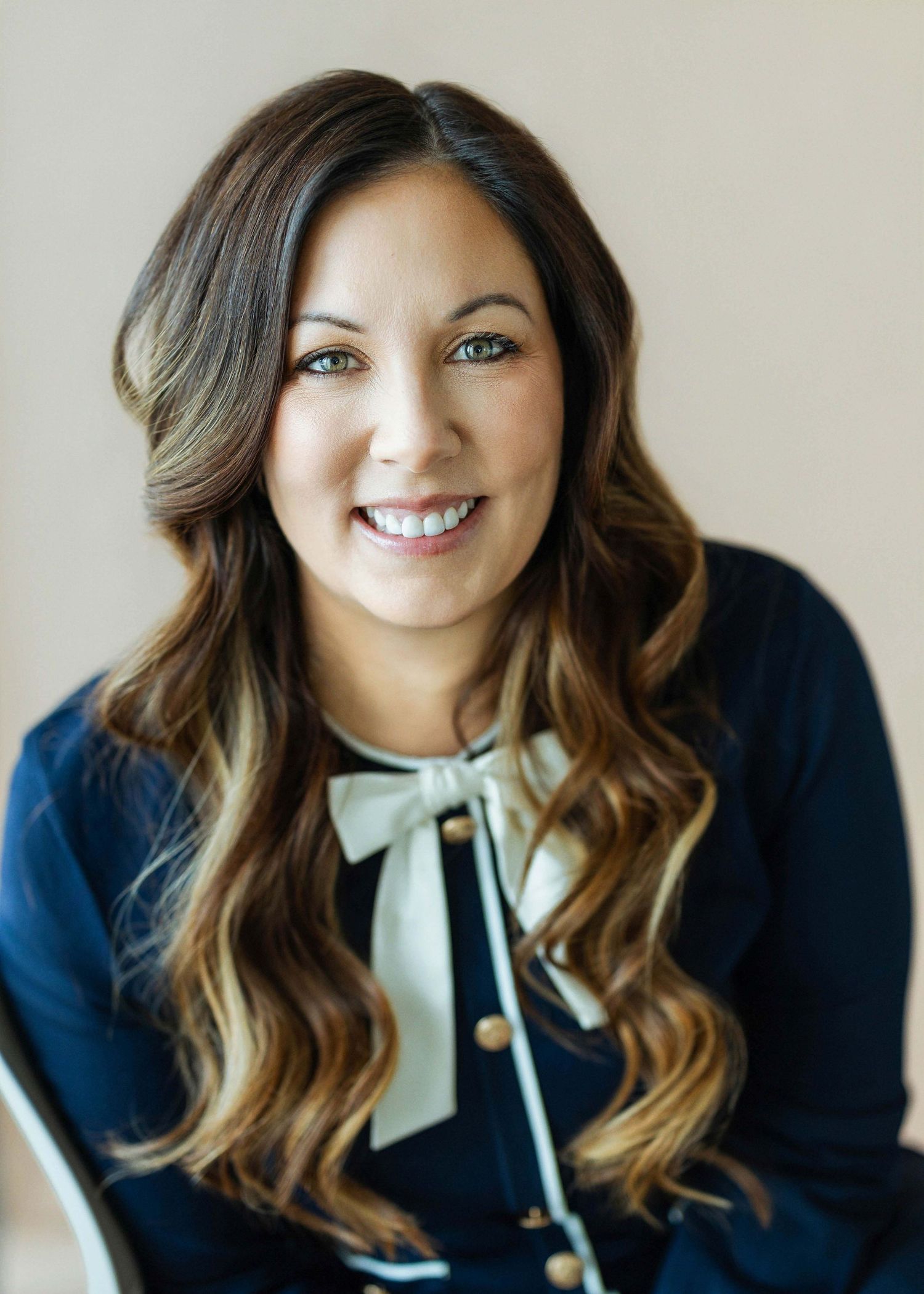 Professional headshot of a smiling woman with long brown highlighted hair wearing a navy blue blouse with bow detail.