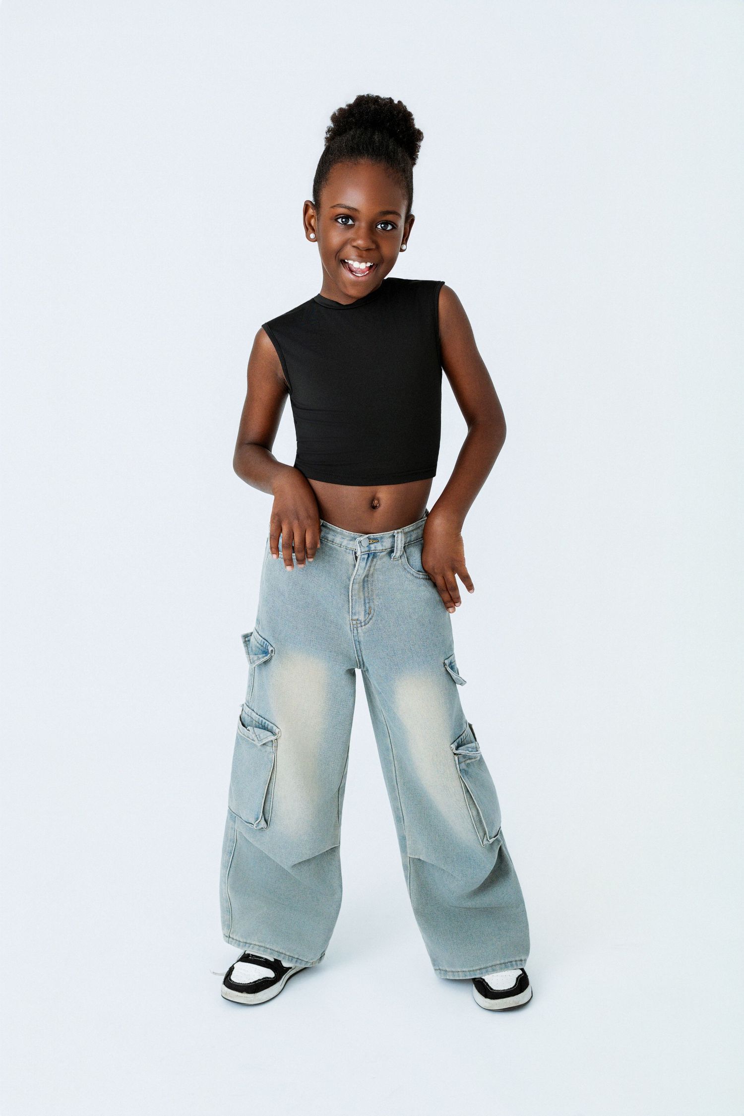 A young girl wearing a black crop top and wide-leg cargo jeans with sneakers, smiling against a white background.