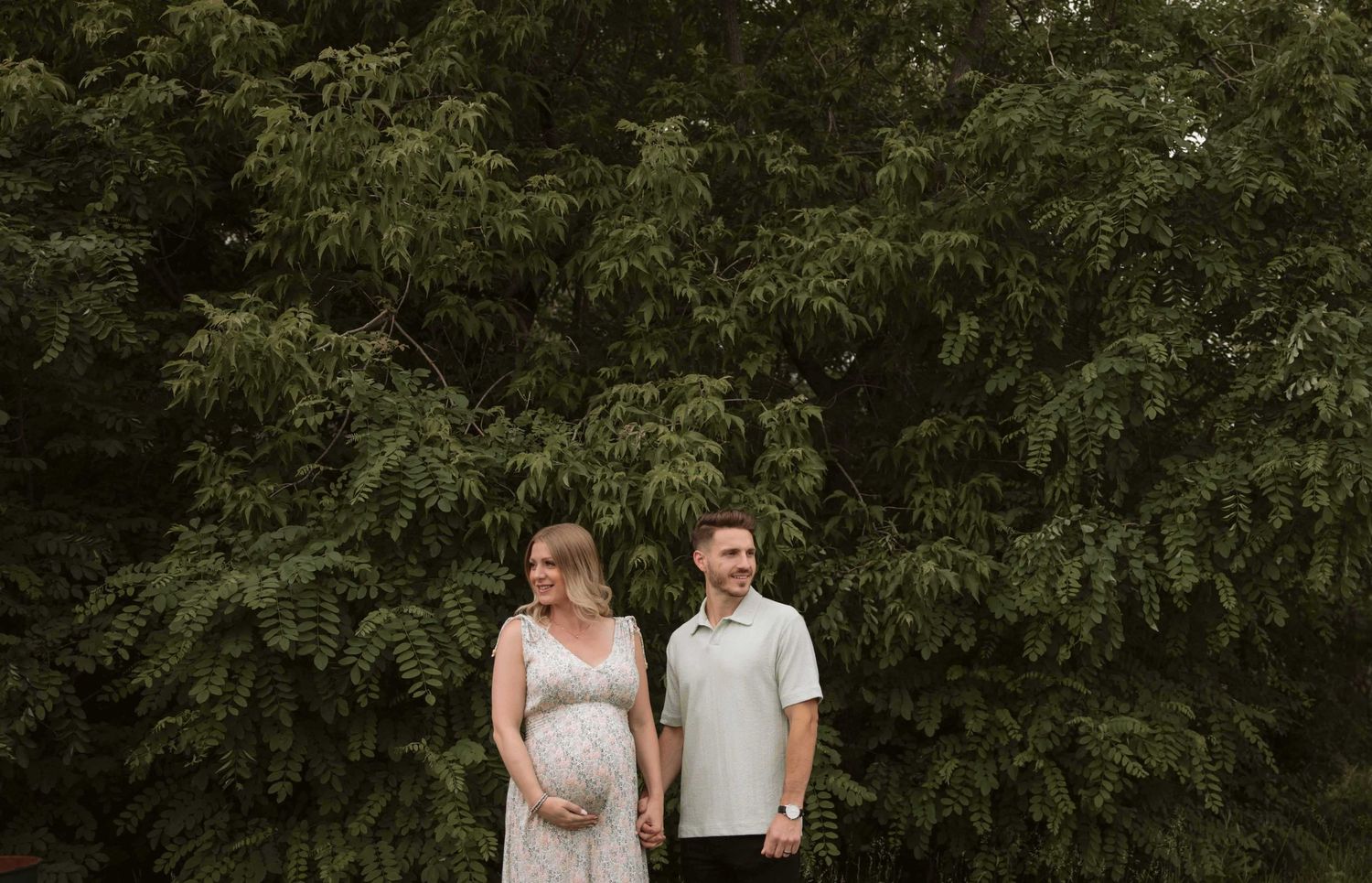 Couple standing together in front of lush green trees during a summer photoshoot.