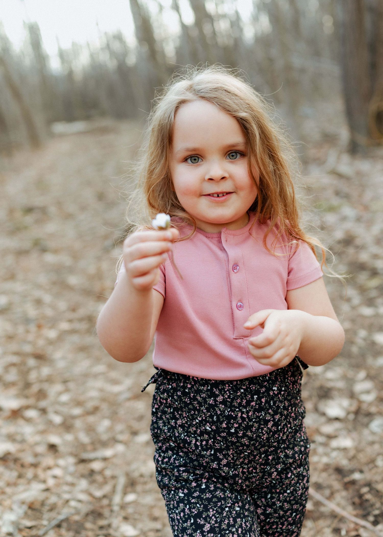 Smiling toddler girl with blonde hair holding a flower in a woodland setting, wearing a pink top and floral pants.