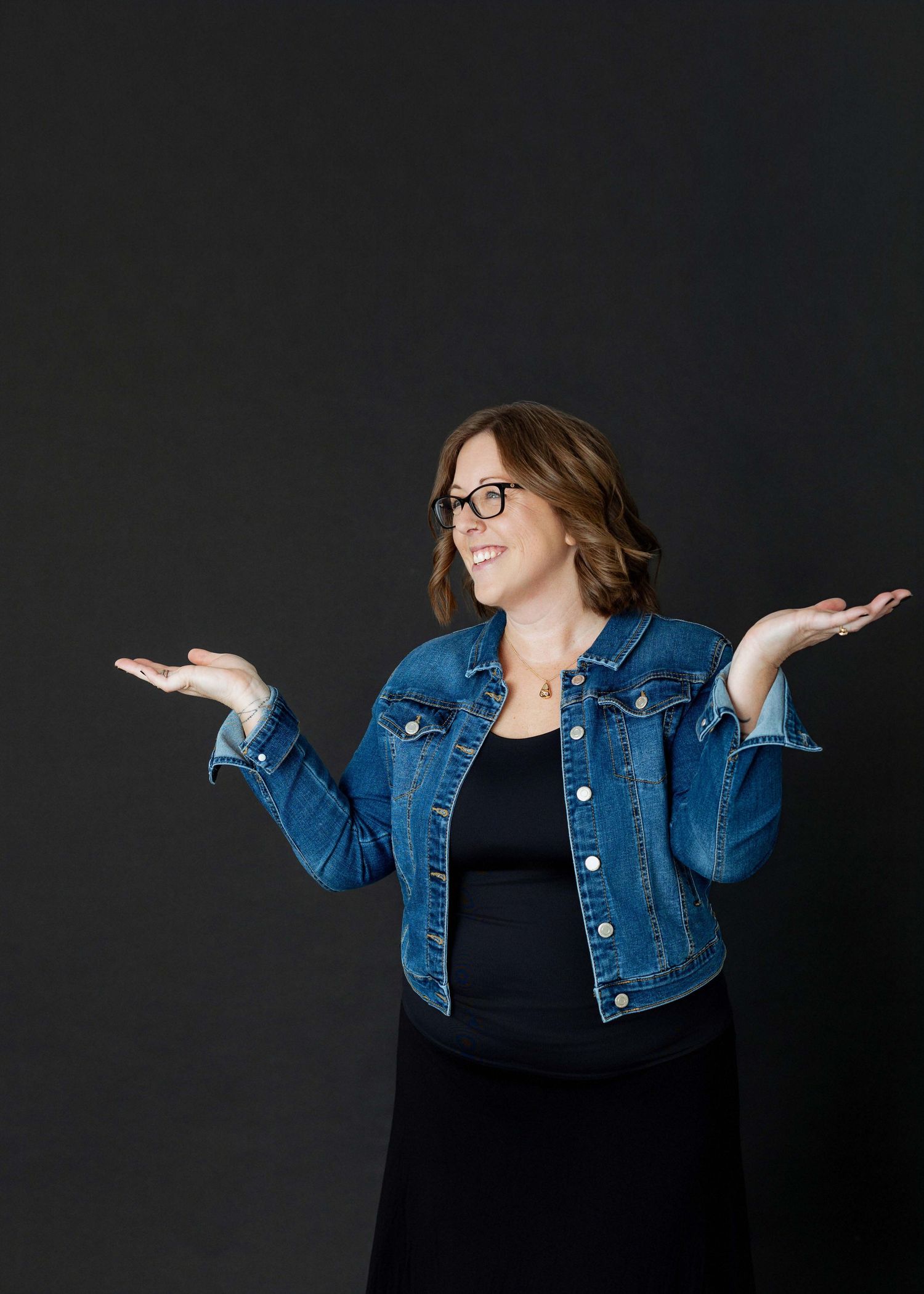 Woman in denim jacket and glasses smiling with hands raised in a shrug pose against dark background.