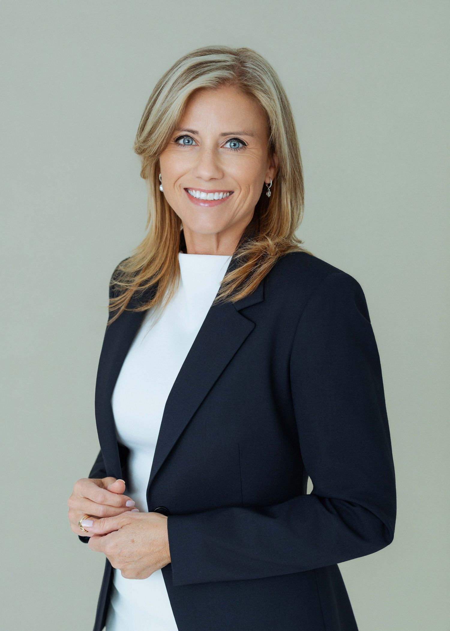 Professional woman in black blazer and white top smiling confidently against a neutral green-gray background.