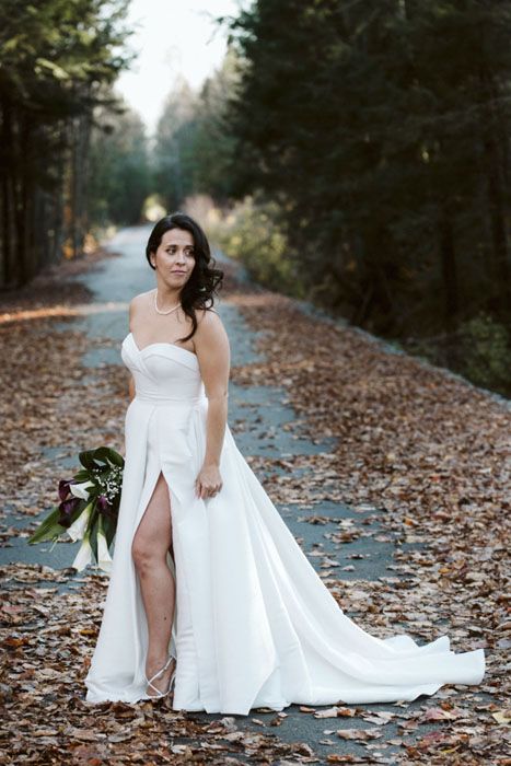 Bride in elegant white gown poses on leaf-covered path during autumn wedding photoshoot.