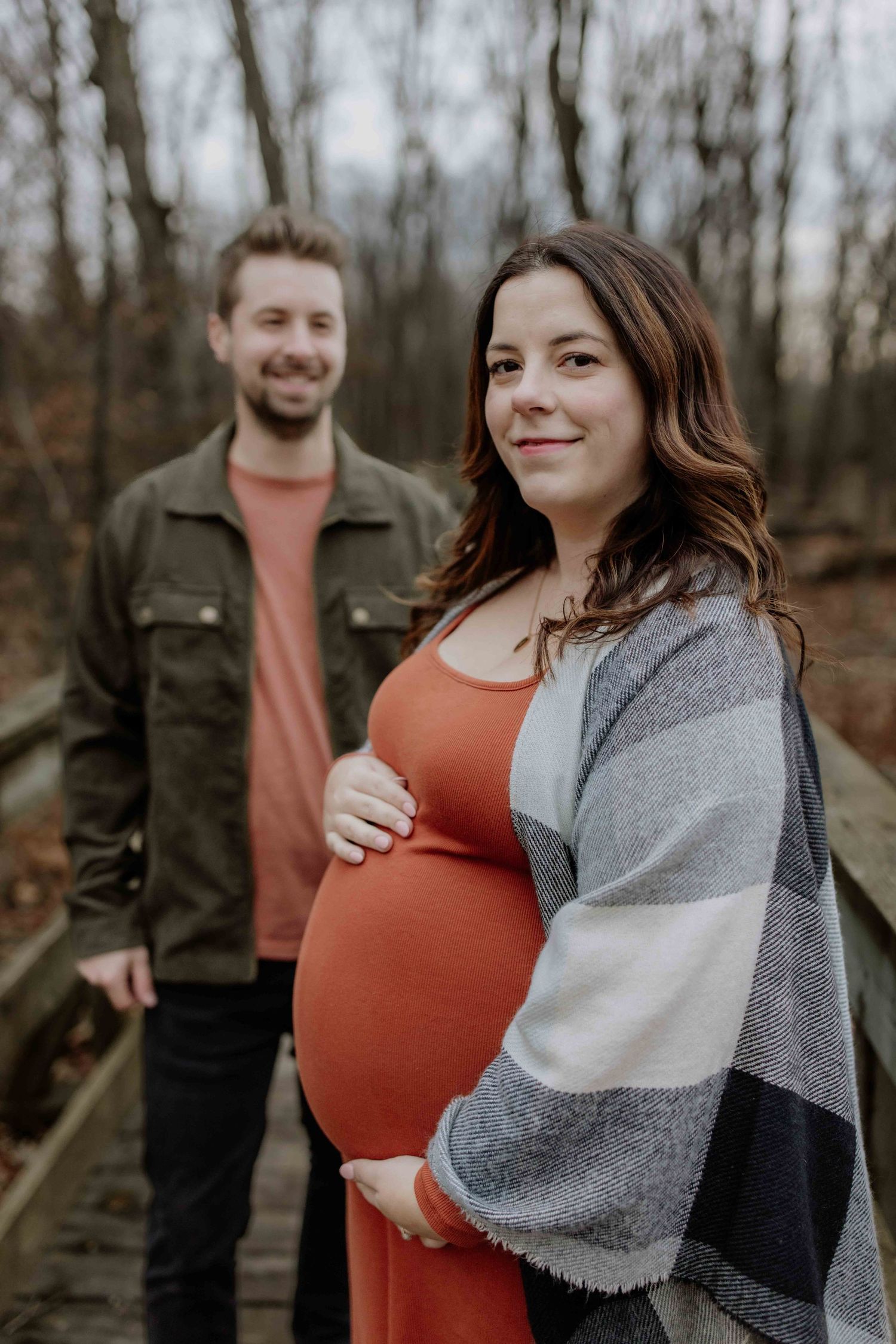 Couple poses on wooden boardwalk during maternity photoshoot in autumn forest setting.