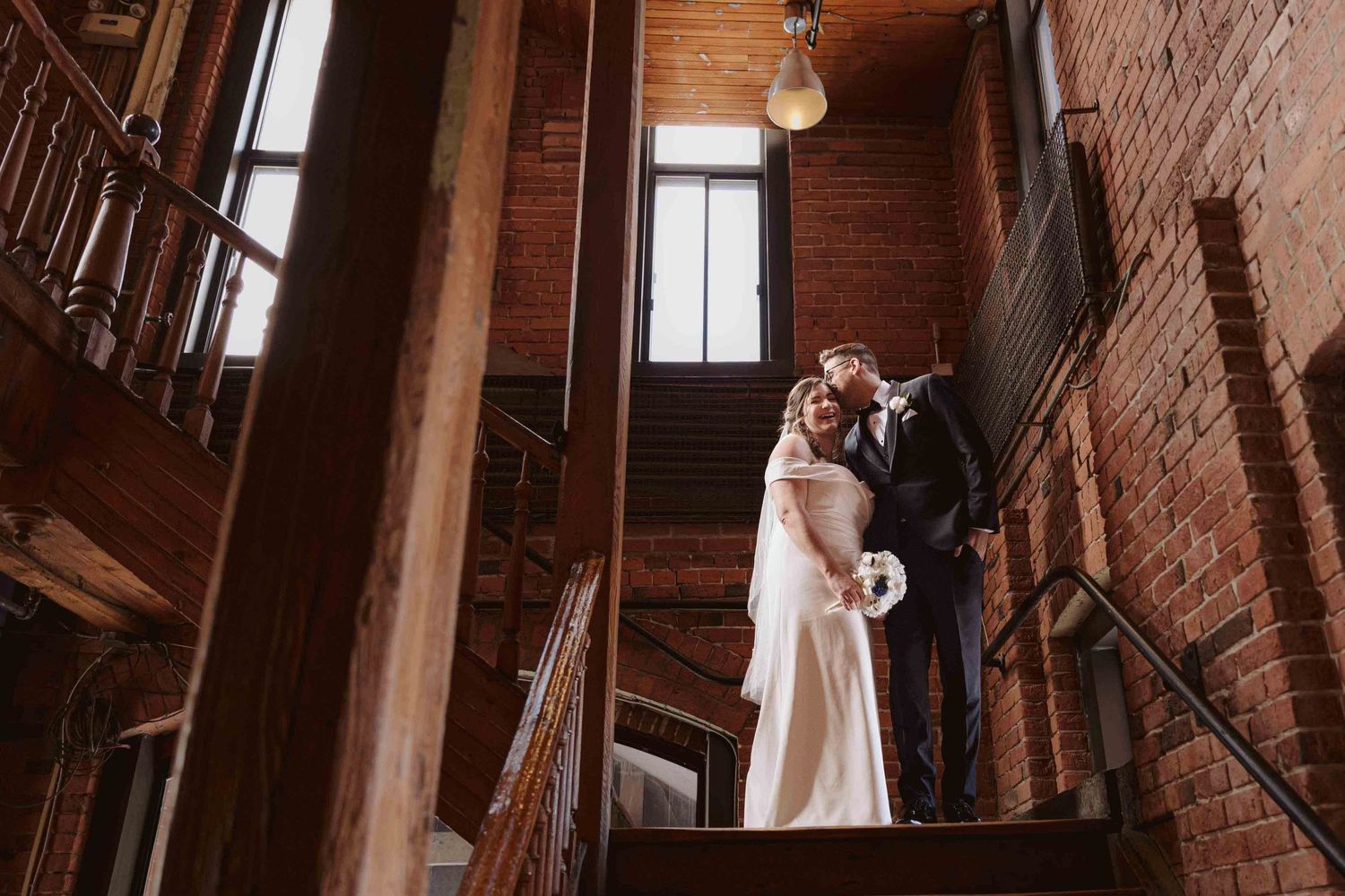 A couple in wedding attire poses in an industrial brick loft space with dramatic window lighting.