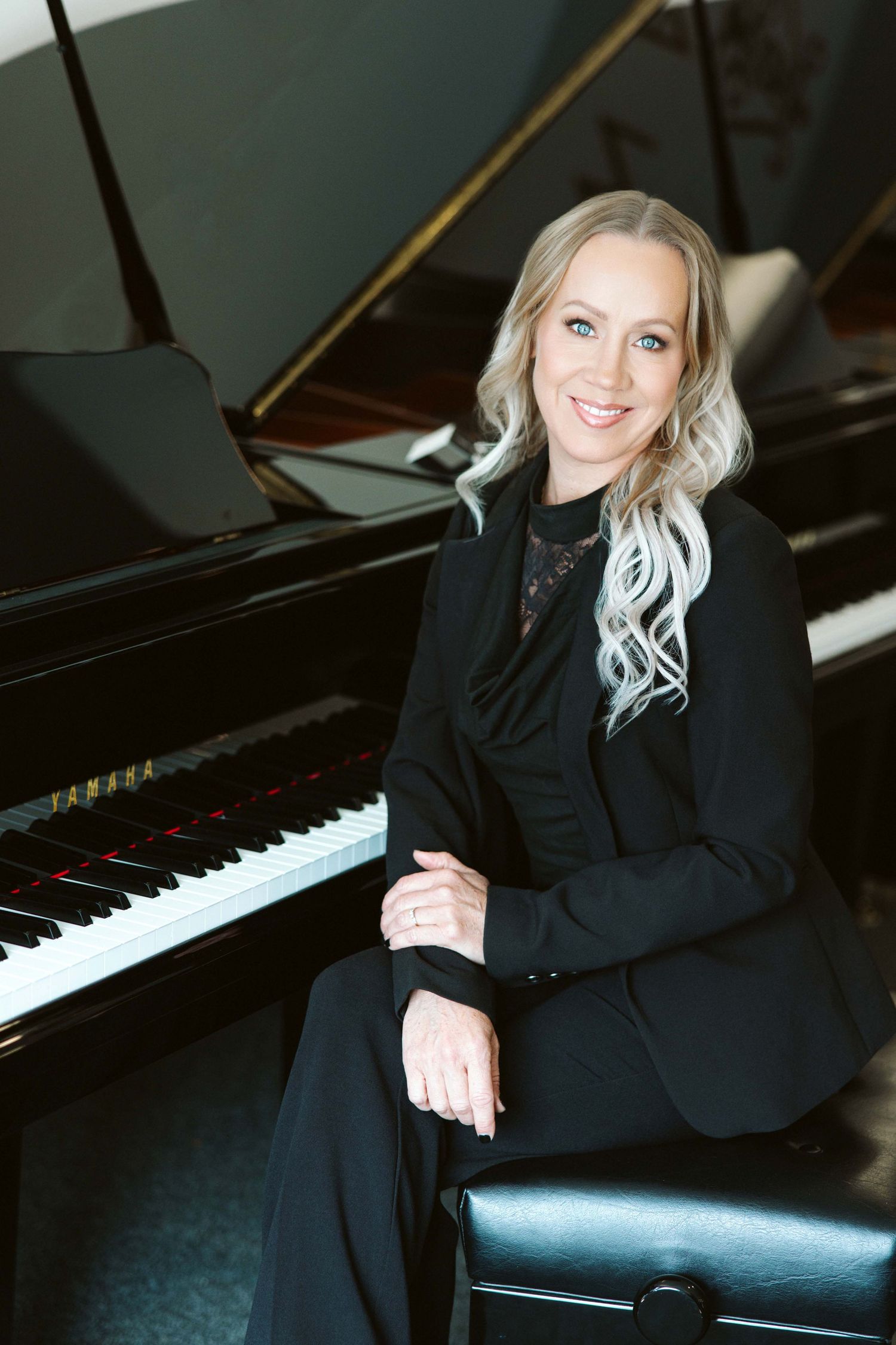 Smiling blonde woman in black suit seated at a Yamaha grand piano in an elegant music studio setting.