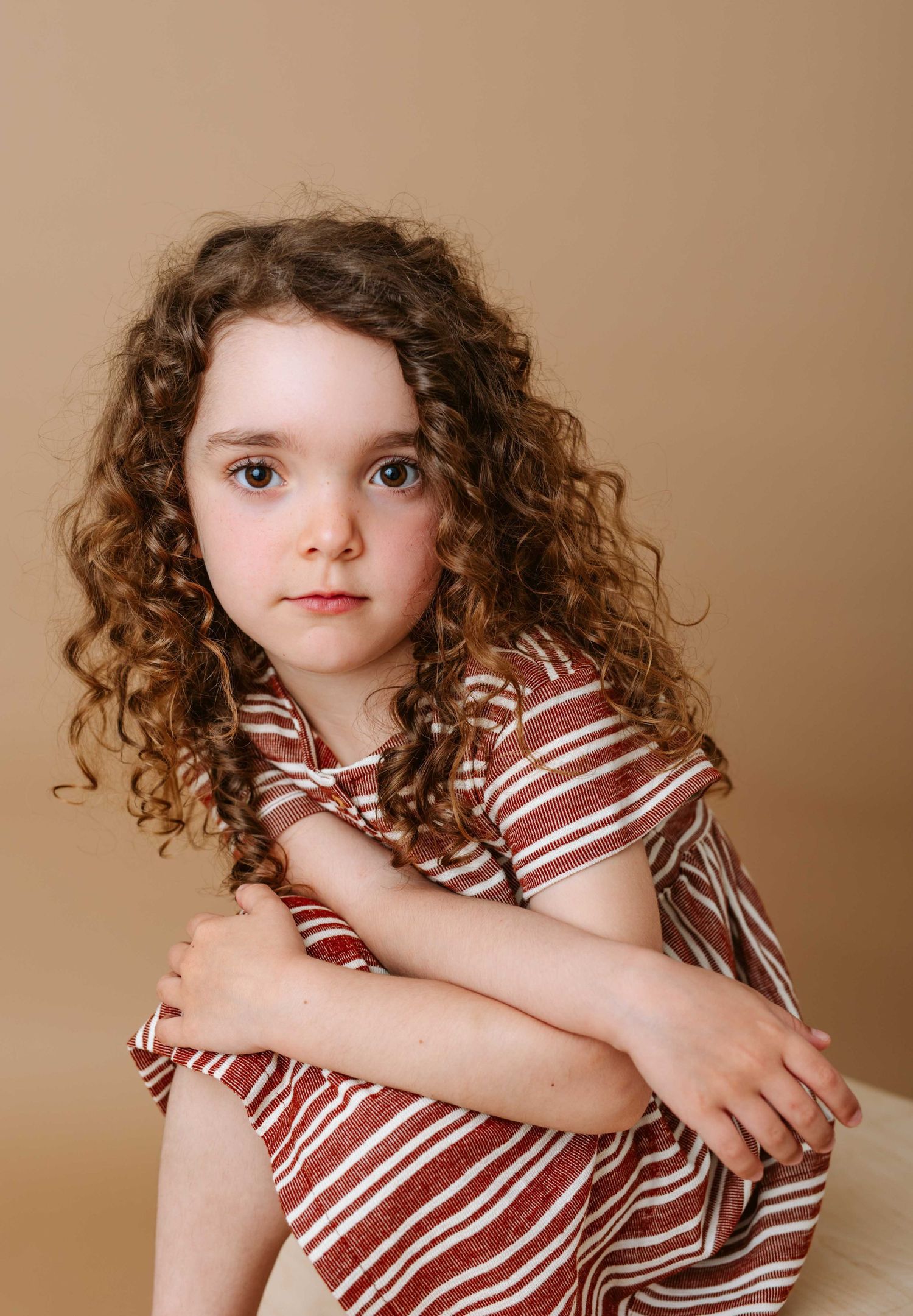 Child in striped dress poses expressively against warm brown studio background.