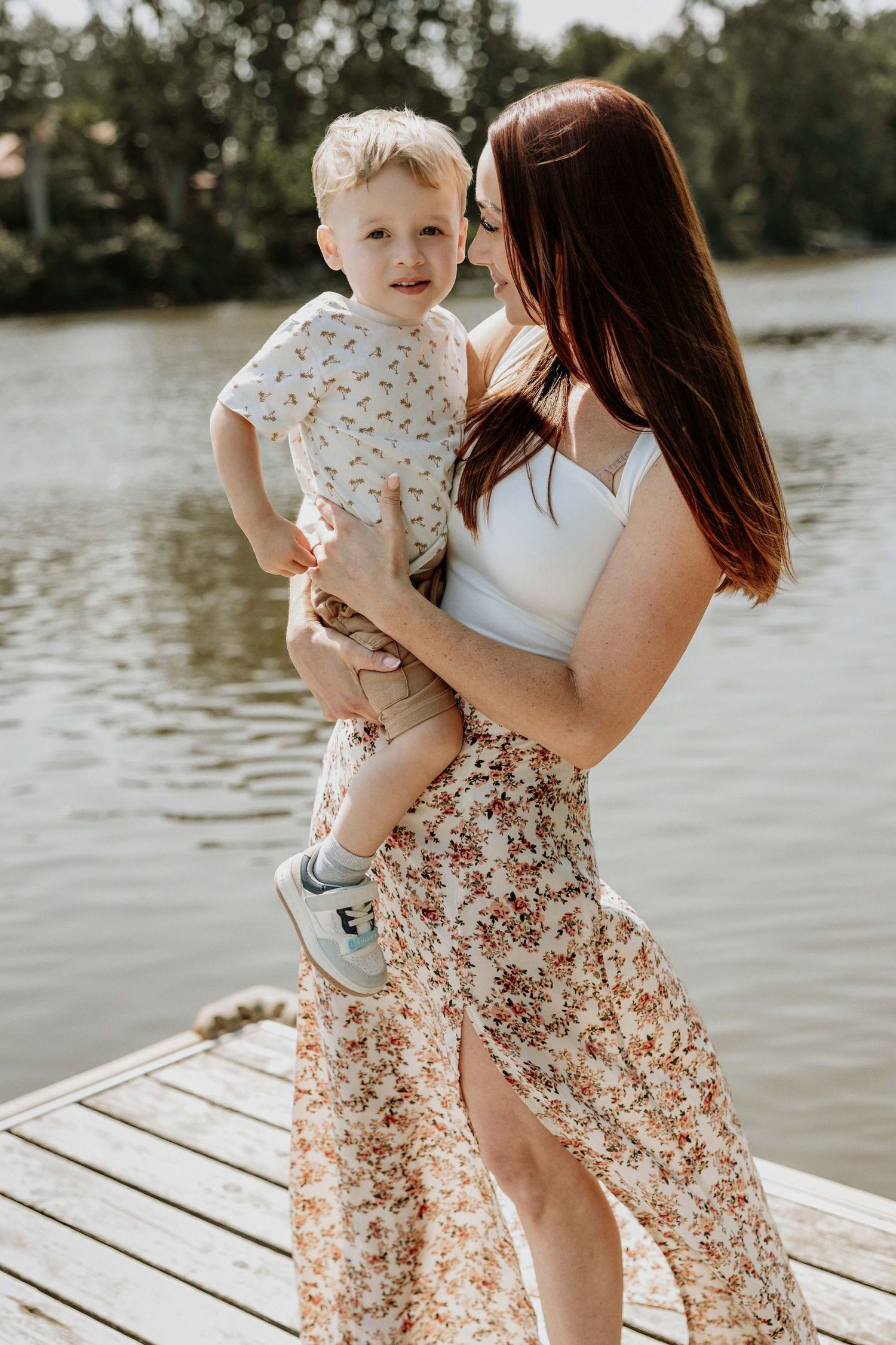 A mother holds her toddler son on a wooden dock by a lake, both wearing floral outfits on a sunny day.