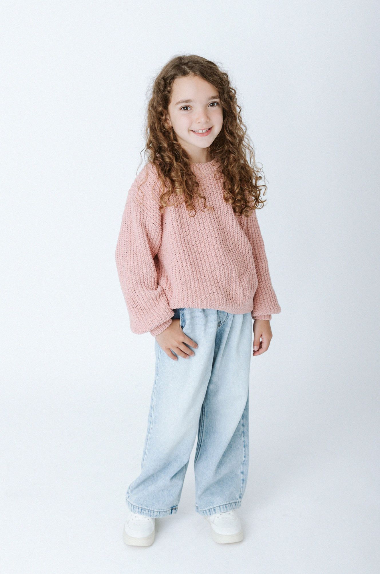 Child wearing pink sweater and light wash jeans poses against white background in casual fashion photo series.