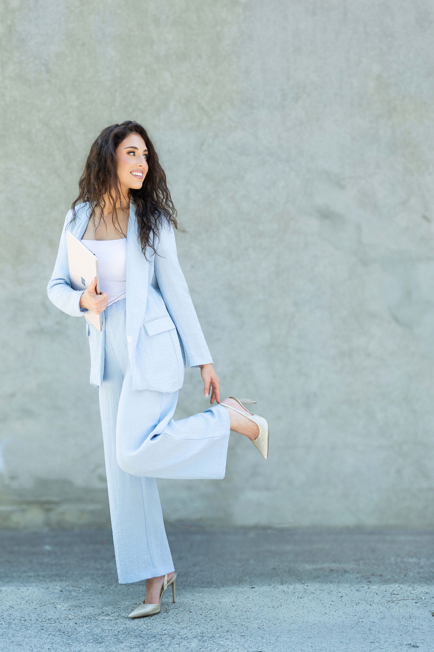Smiling woman in light blue pantsuit holding a folder, posing outdoors against a gray concrete wall. corporate