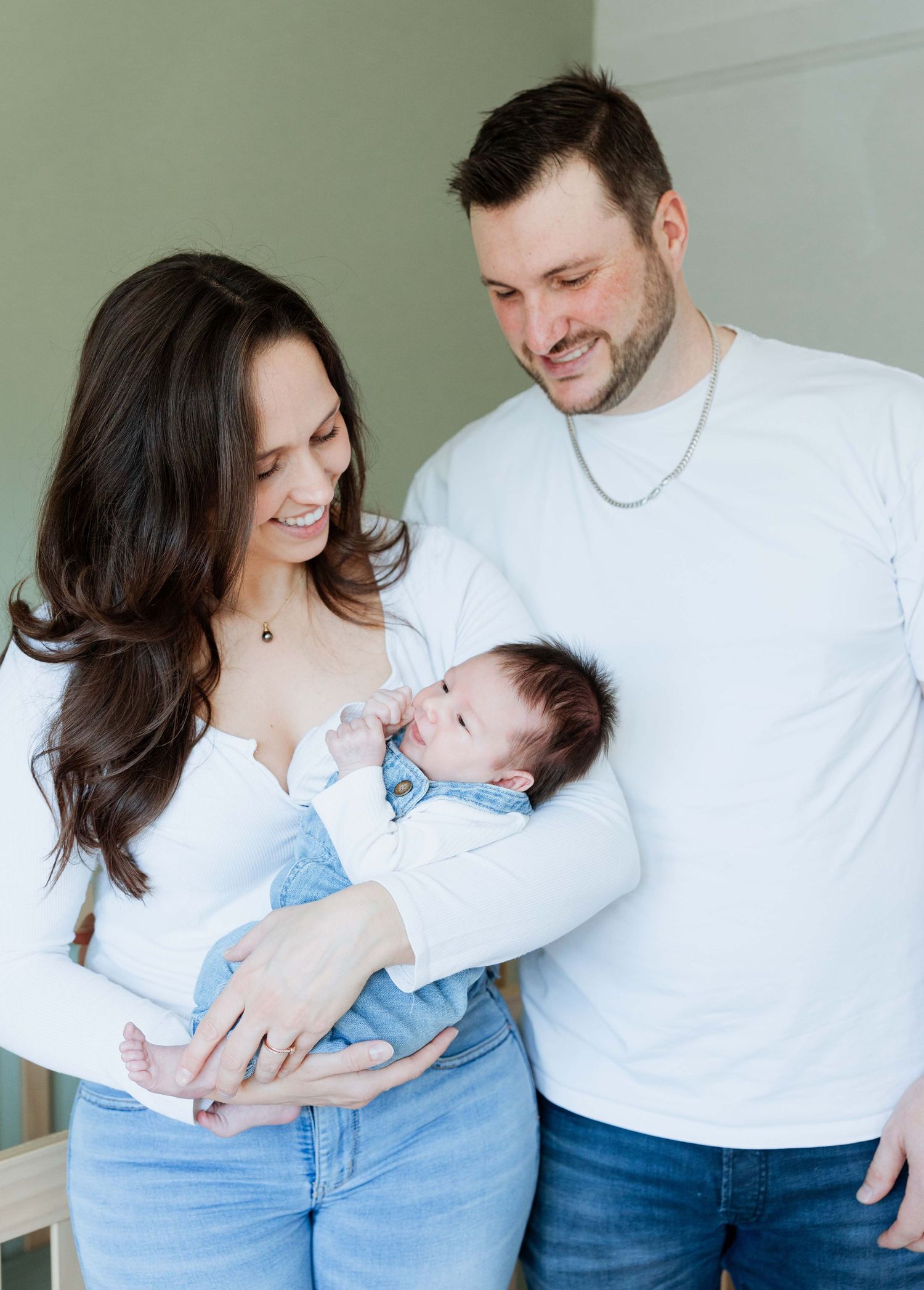 A smiling couple in white tops and jeans lovingly gazes at their newborn baby during a family portrait session.