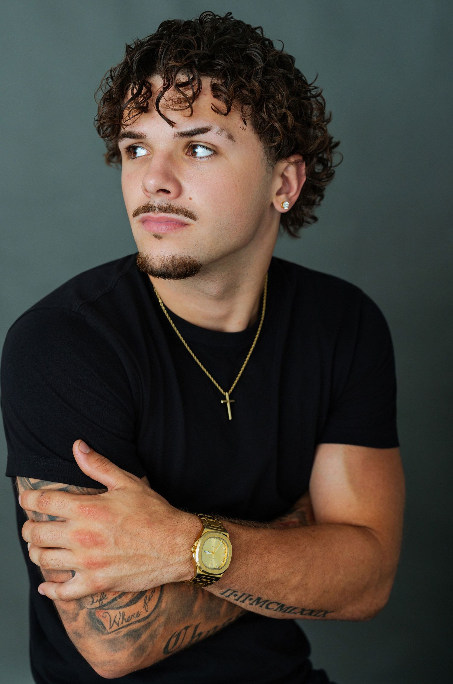 Young man with curly hair, tattoos, gold chain necklace, and watch posing against a gray background.